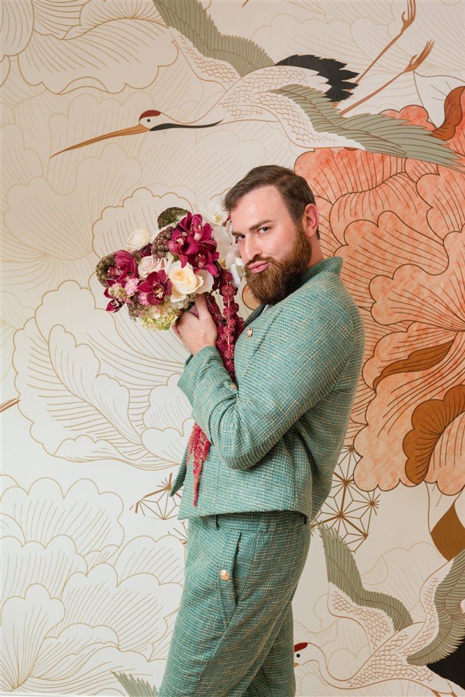 The Bridal Steward holding a bouquet of flowers, standing in front of a decorative wall with crane and floral patterns.