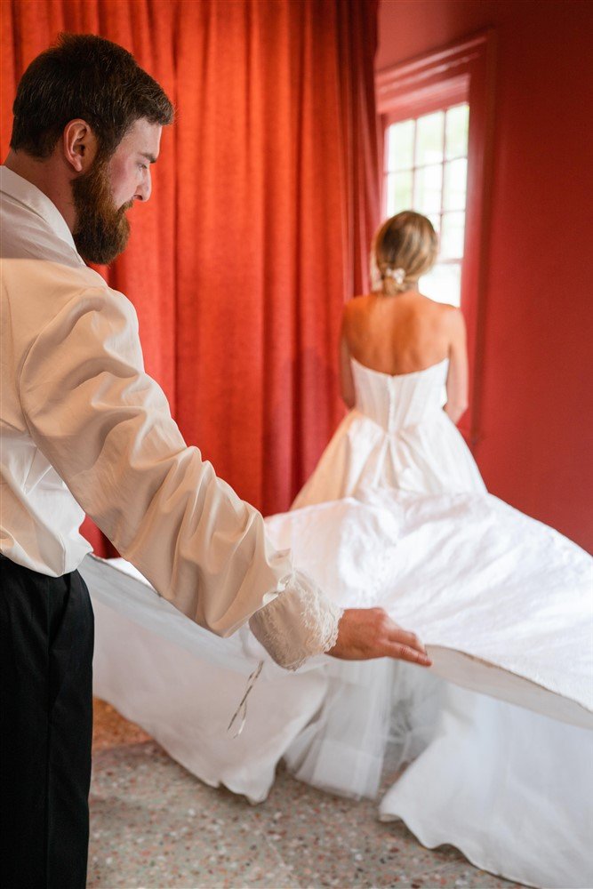 The Bridal Steward adjusting a wedding dress, which is displayed on a bride, in a wedding suite with red curtains and a window in the background.