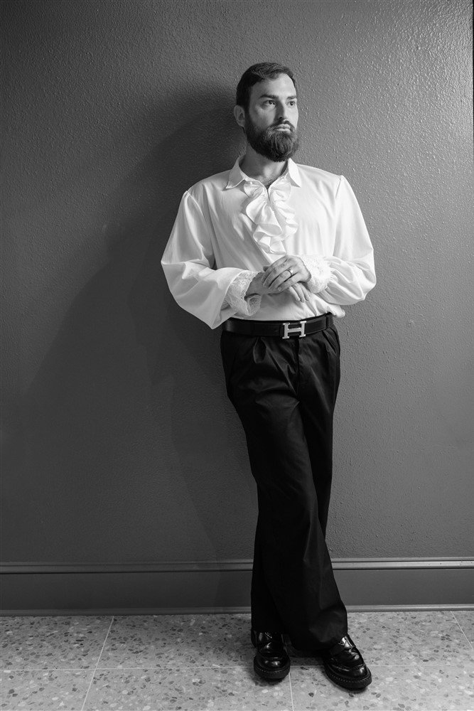 A black and white photo of The Bridal Steward, standing against a textured wall. He is wearing a white ruffled shirt, black trousers with an 'H' belt, and shiny black shoes. He has folded his hands in front and is looking to the side.