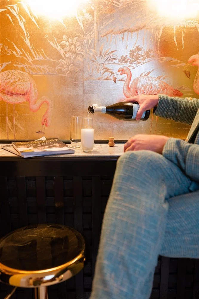 The Bridal Steward in a plaid suit is sitting at a bar, pouring champagne into a glass. The background features a mural of pink flamingos and tropical plants.
