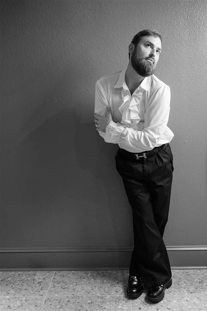 The Bridal Steward leaning against a textured wall, wearing a white ruffled shirt, black pants, and shiny black shoes, with arms crossed and a contemplative expression.