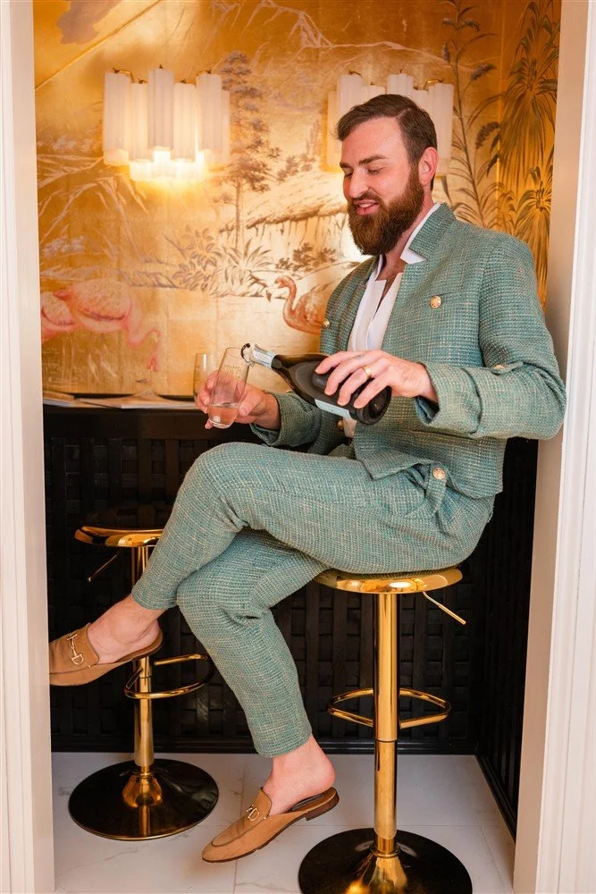 The Bridal Steward in a green plaid suit sitting on a gold bar stool, pouring a drink from a bottle into a glass, in a decorated room with mural wallpaper of landscape and swans, and a wall-mounted light fixture.