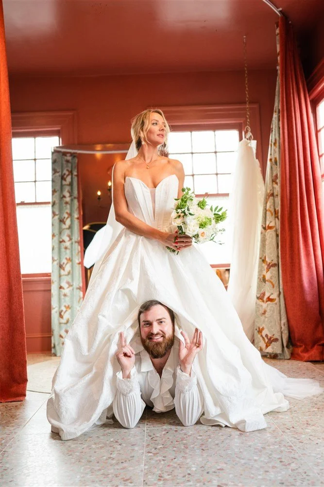 Bride standing over The Bridal Steward inside a room with red walls and curtains, holding a bouquet, with windows and curtains in the background.