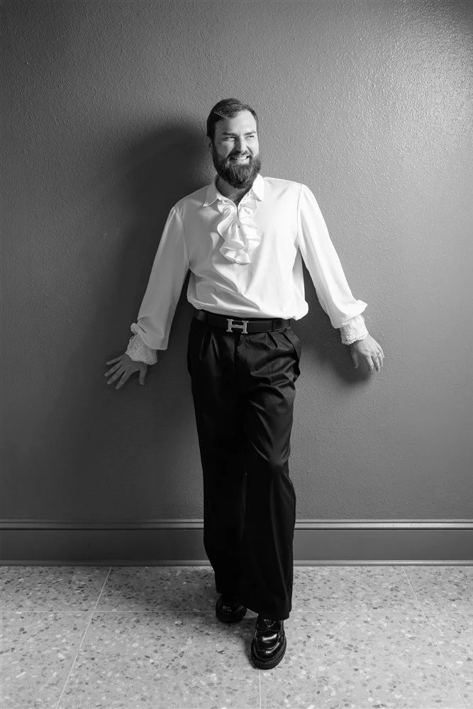 Black and white photo of a The Bridal Steward standing against a wall, smiling, wearing a white shirt with ruffled cuffs and a front ruffle, black belt, and black pants.
