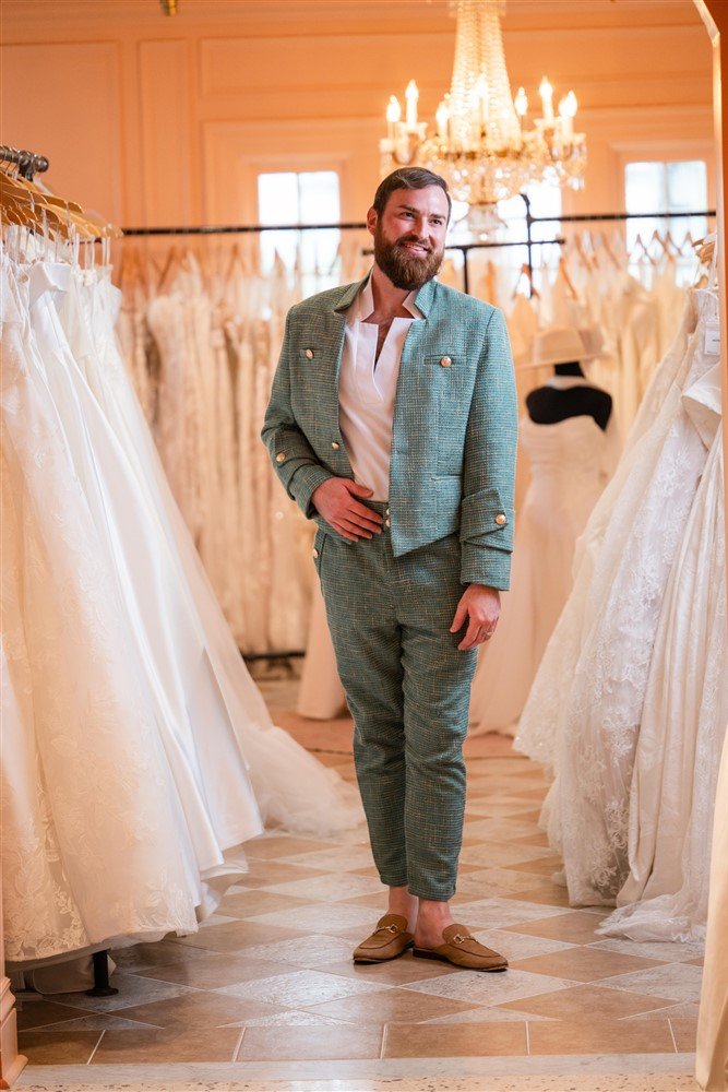 The Bridal Steward wearing a green suit and loafers, smiling, standing inside a wedding dress boutique surrounded by wedding gowns and a chandelier.