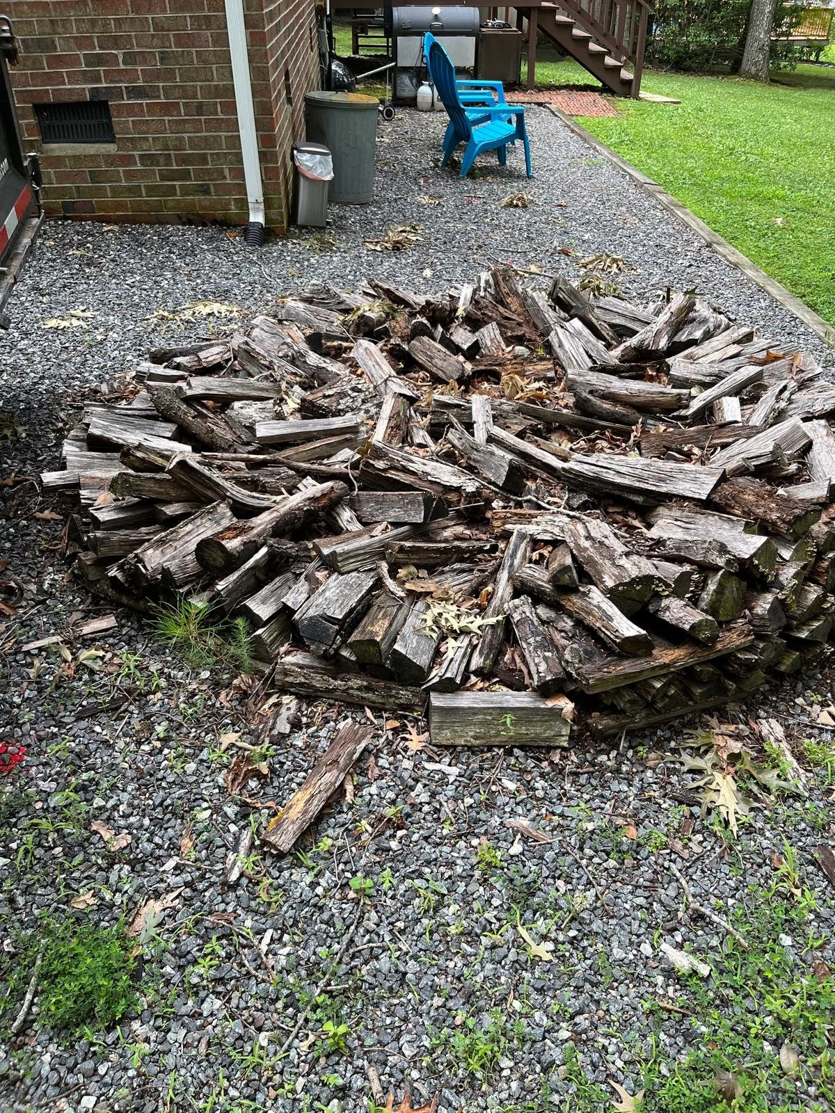 A pile of cut firewood logs on a gravel backyard surface near a brick house wall with a trash can and an outdoor chair, with a grassy yard and trees in the background.