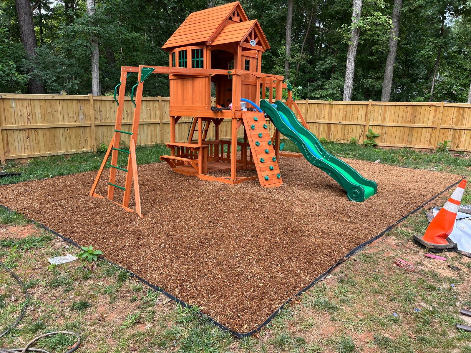 A new wooden playset with a slide, swings, and a climbing wall in a backyard, surrounded by a wooden fence and trees.
