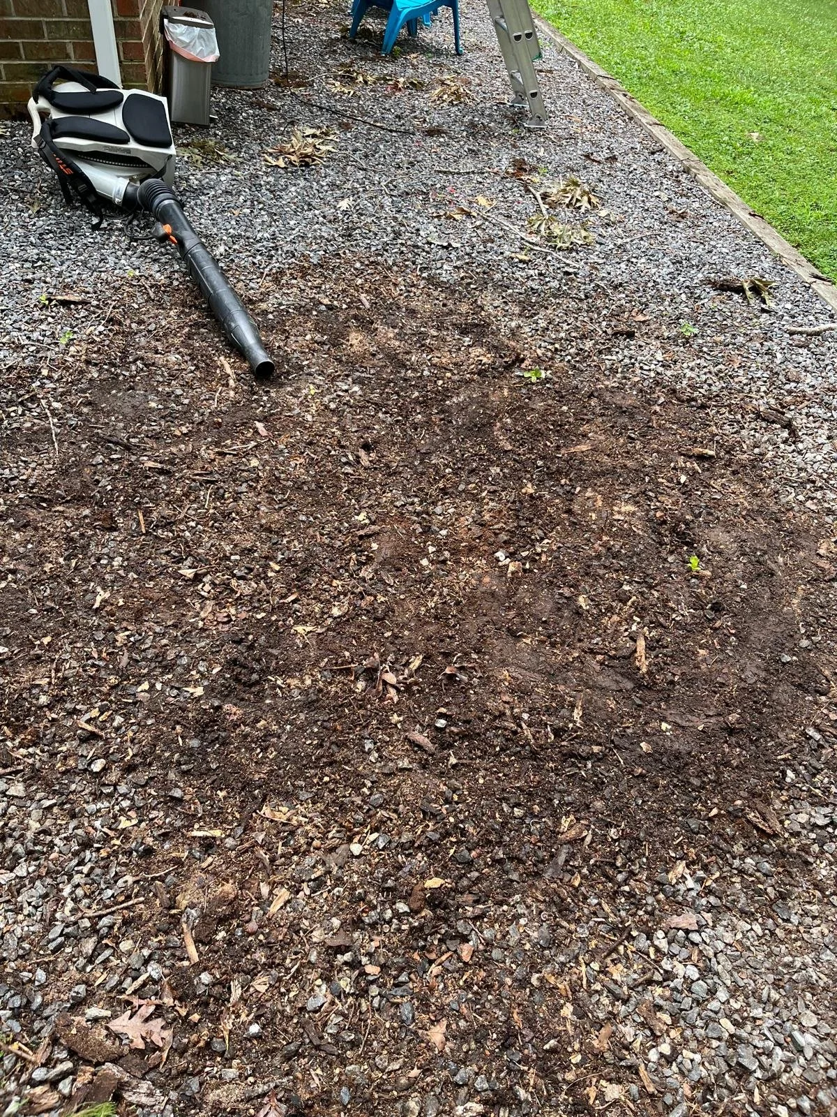 A backyard garden area with a section of soil prepared for planting. There is a leaf blower lying on the ground, a trash can, a ladder, and some outdoor chairs in the background.