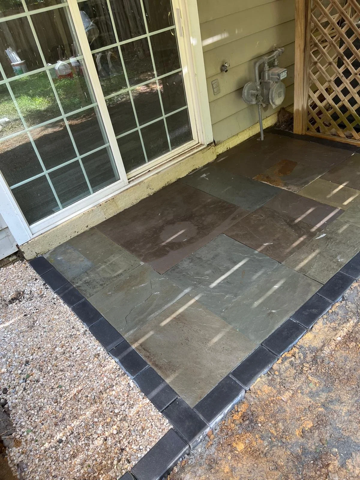 Exterior patio with large stone tiles, a sliding glass door, and an outdoor gas meter with pipes. Gravel surrounds the tiled area.