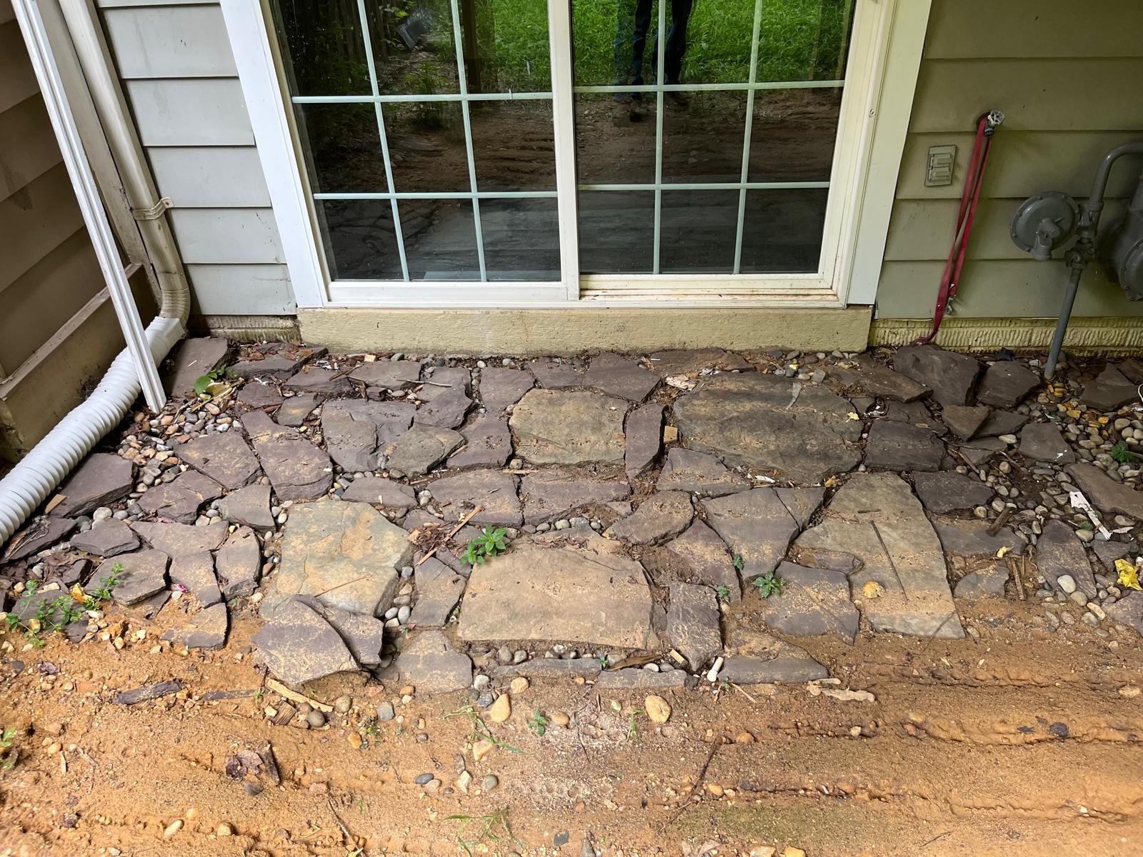 A stone pathway outside a house with a sliding glass door. The pathway is made of large, flat stones surrounded by gravel and small plants. The house exterior is painted light green with horizontal siding. There is a white drainpipe on the left, and 