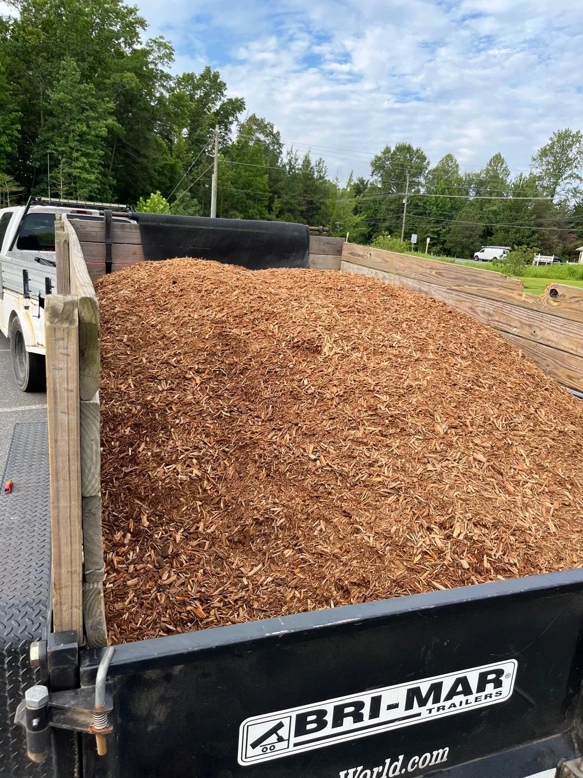 A trailer loaded with a large pile of wood mulch, with a pickup truck and trees in the background and a partly cloudy sky.