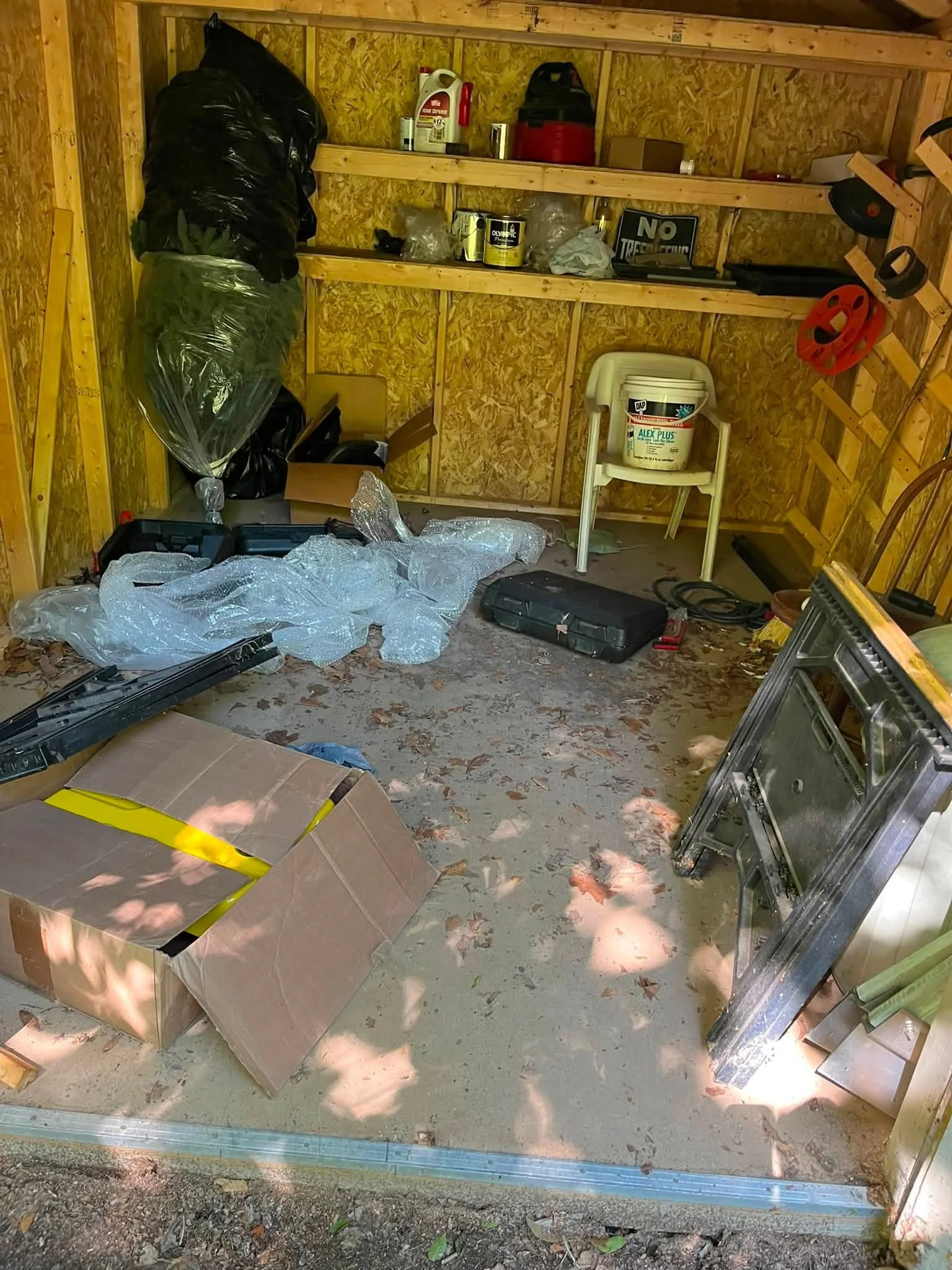 Inside a wooden shed with clutter including cardboard boxes, plastic bags, paint cans, a high chair, and tools on the floor and shelves.