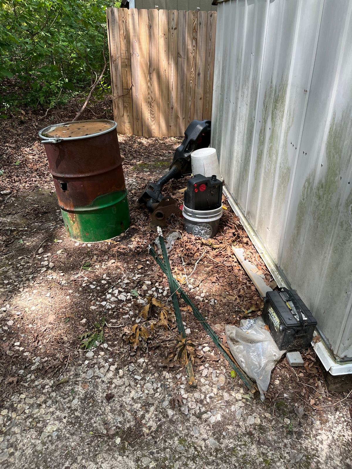 An outdoor area with a rusty barrel, a black cordless leaf blower, a white bucket, a battery, and miscellaneous tools scattered on the ground among fallen leaves and small rocks, next to a metal fence and a wooden fence.