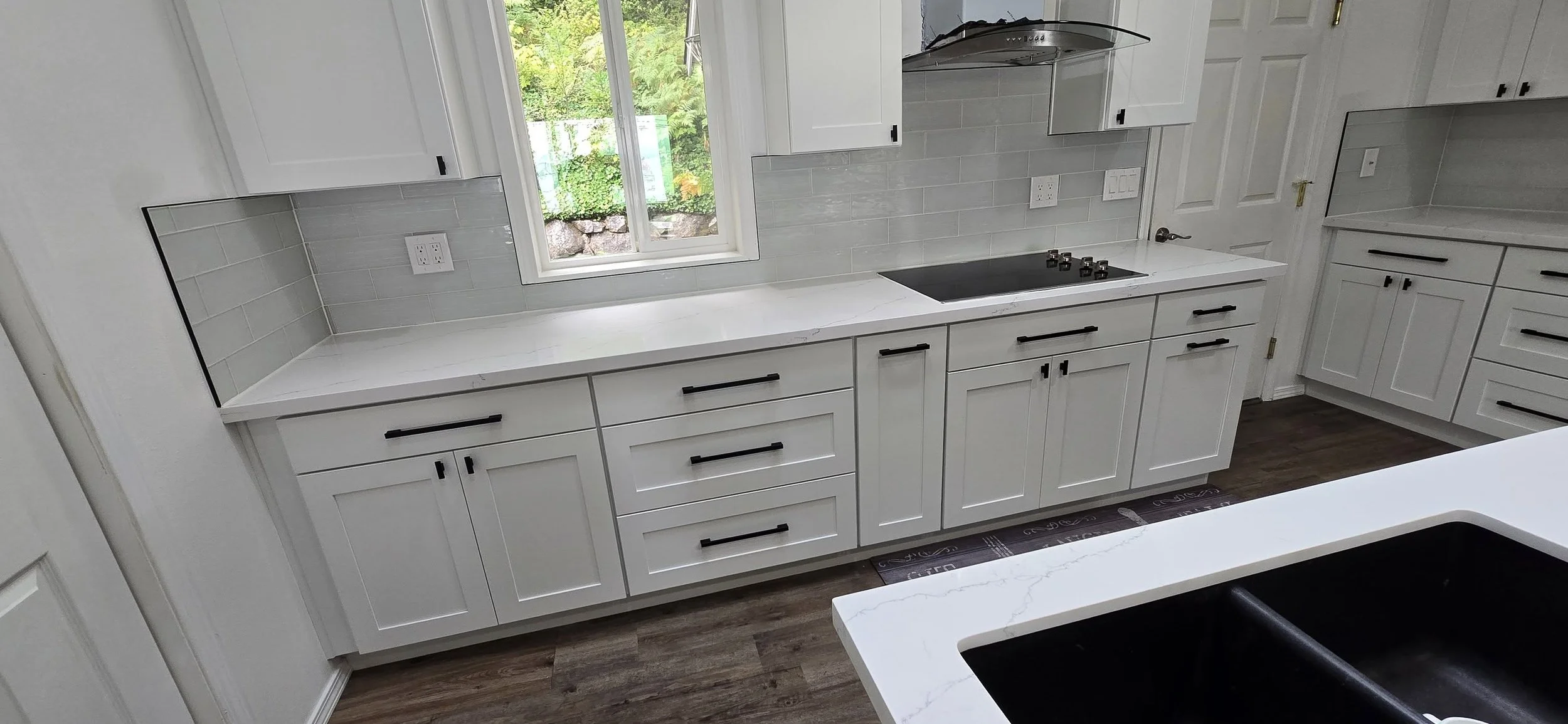 Modern kitchen with white cabinets, black handles, light gray tile backsplash, and a window overlooking greenery.