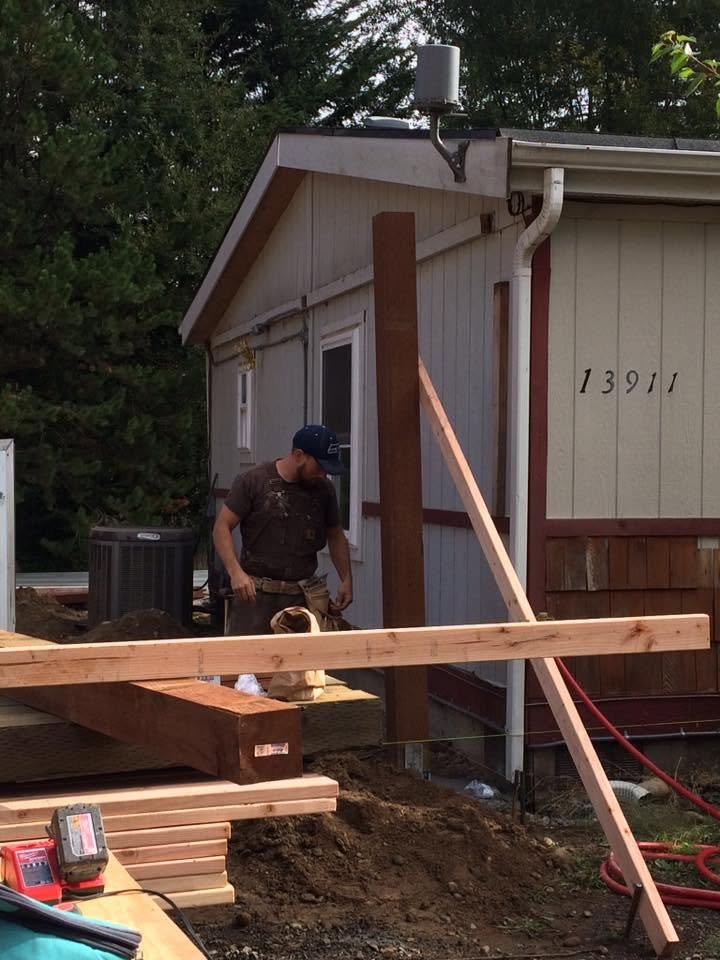 A construction worker inspecting a wooden support post next to a house with the street number 13911. The house has beige and brown exterior siding, a slanted roof, and some construction materials and tools are visible in the yard.