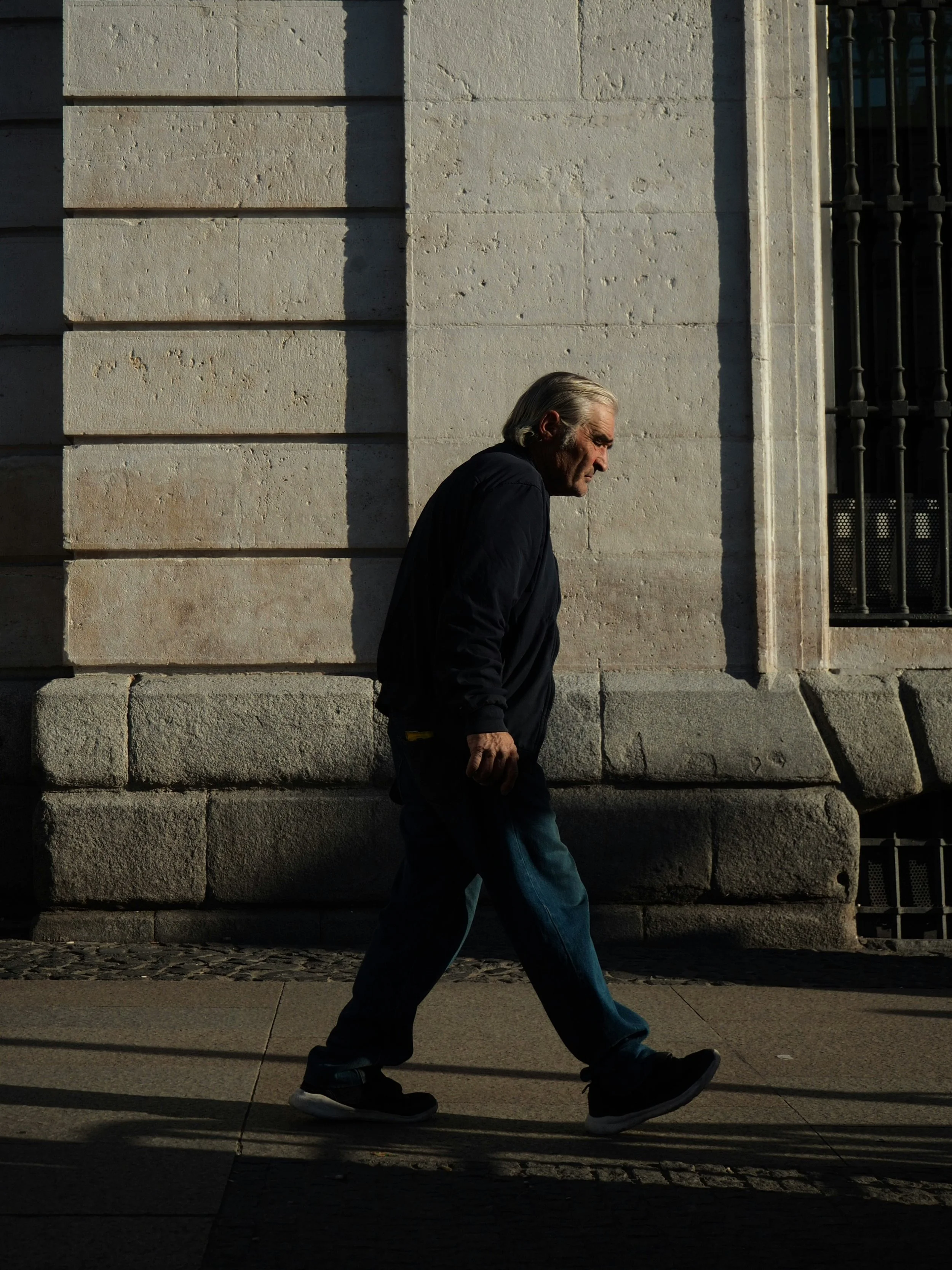 An older man walking on the sidewalk in front of a stone building during daylight.
