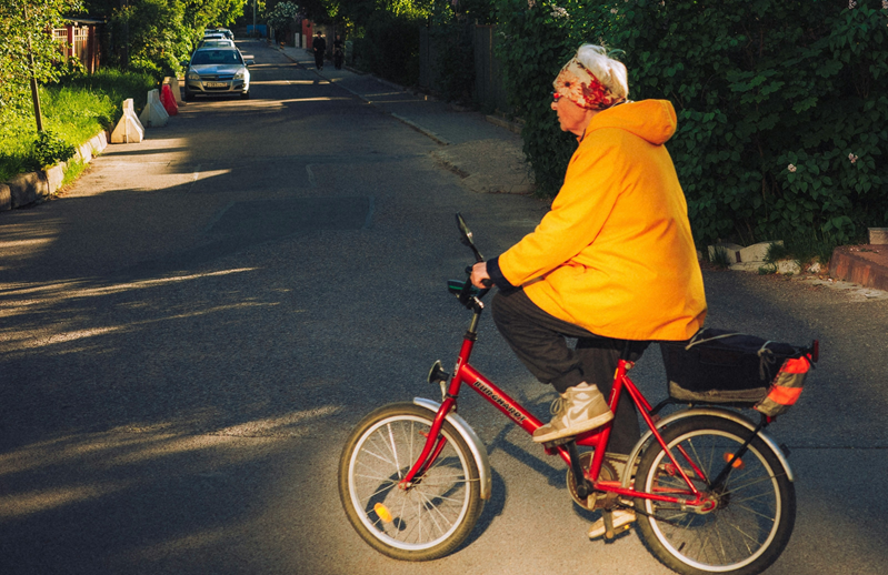 An elderly person riding a red bicycle on a residential street, wearing a yellow jacket and a headband.