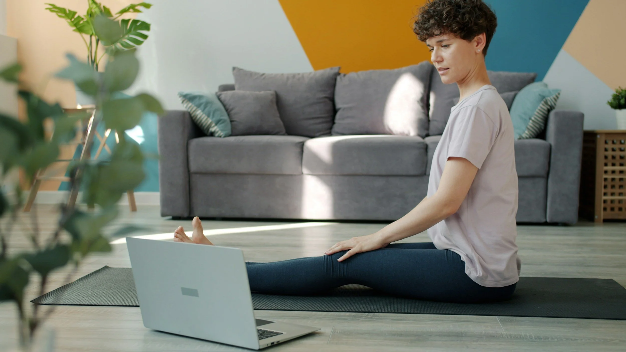 A woman practicing yoga or meditation in a living room, sitting on a yoga mat with her legs crossed, with an open laptop in front of her, and a gray couch with pillows in the background.