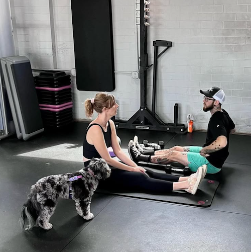 A woman, a man, and a dog in a gym. The woman and the man are sitting on workout mats, facing each other with their legs extended. The woman is smiling and talking, while the man is listening and also smiling. The dog is sitting next to the woman, looking at her.