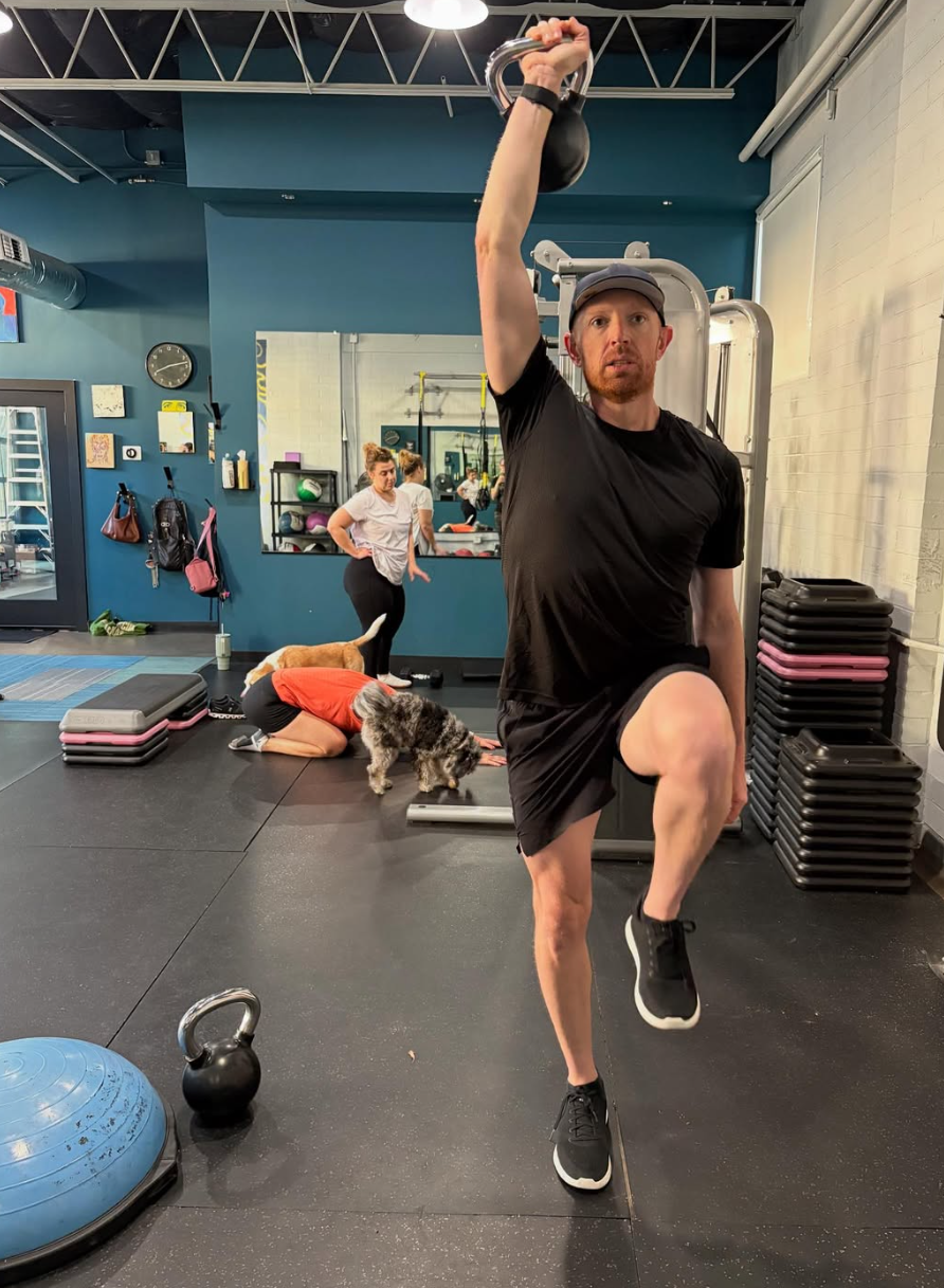 Man lifting a kettlebell with one arm in a gym, with a woman and a dog in the background.