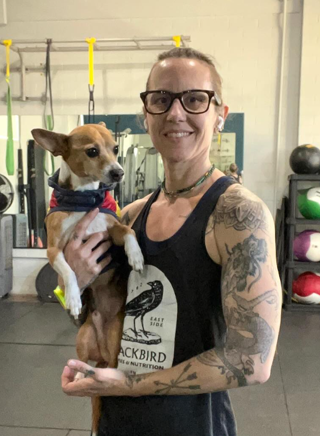 A woman with glasses and tattoos holding a small brown and white dog in a gym. The background shows gym equipment and medicine balls.
