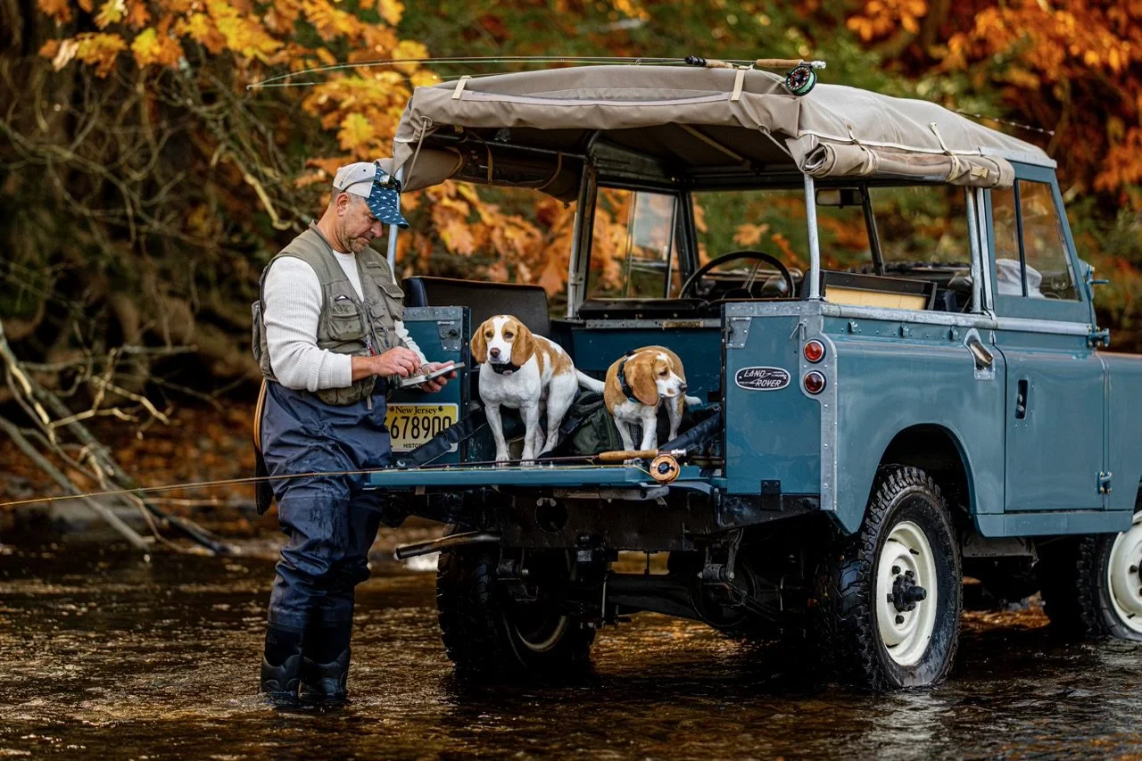 A man standing in a shallow river next to a blue Land Rover with two dogs on the back. The man is writing on a notepad, and the dogs are sitting on a pack in the back of the vehicle, with autumn-colored trees in the background.
