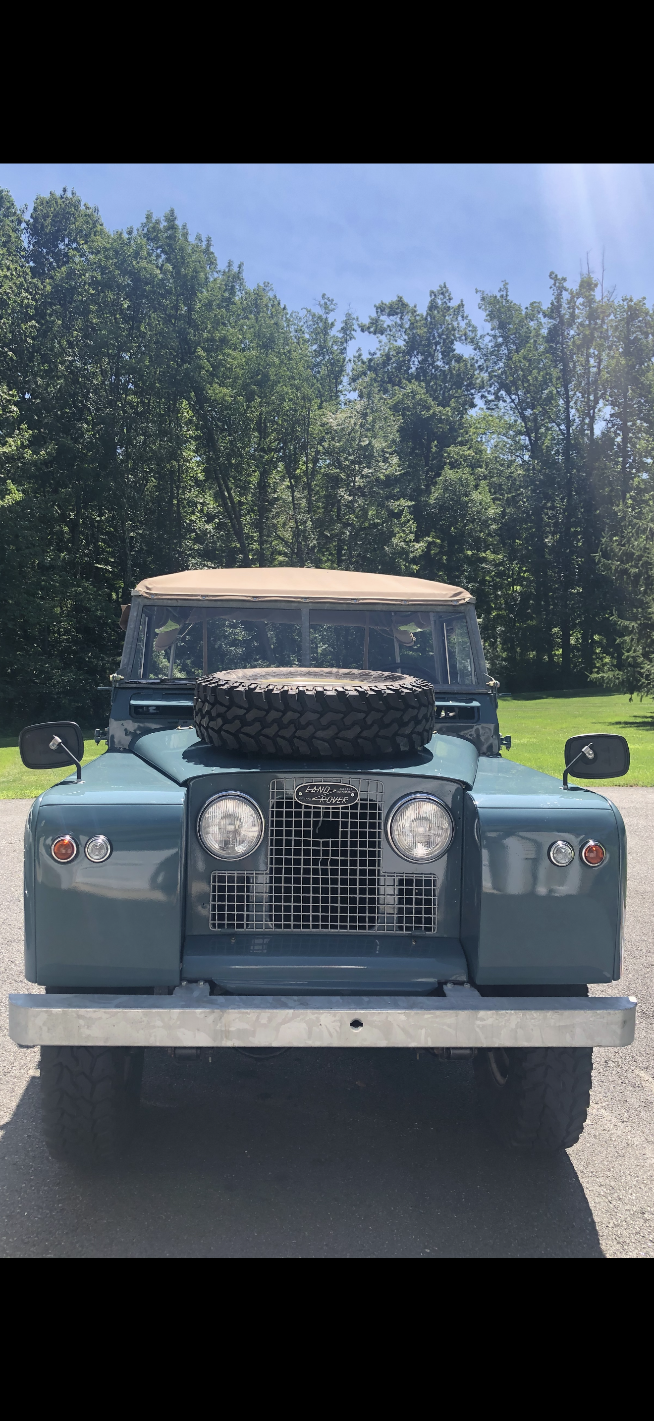 Front view of a vintage blue Land Rover with a spare tire on the hood, set against a backdrop of green trees and a blue sky.