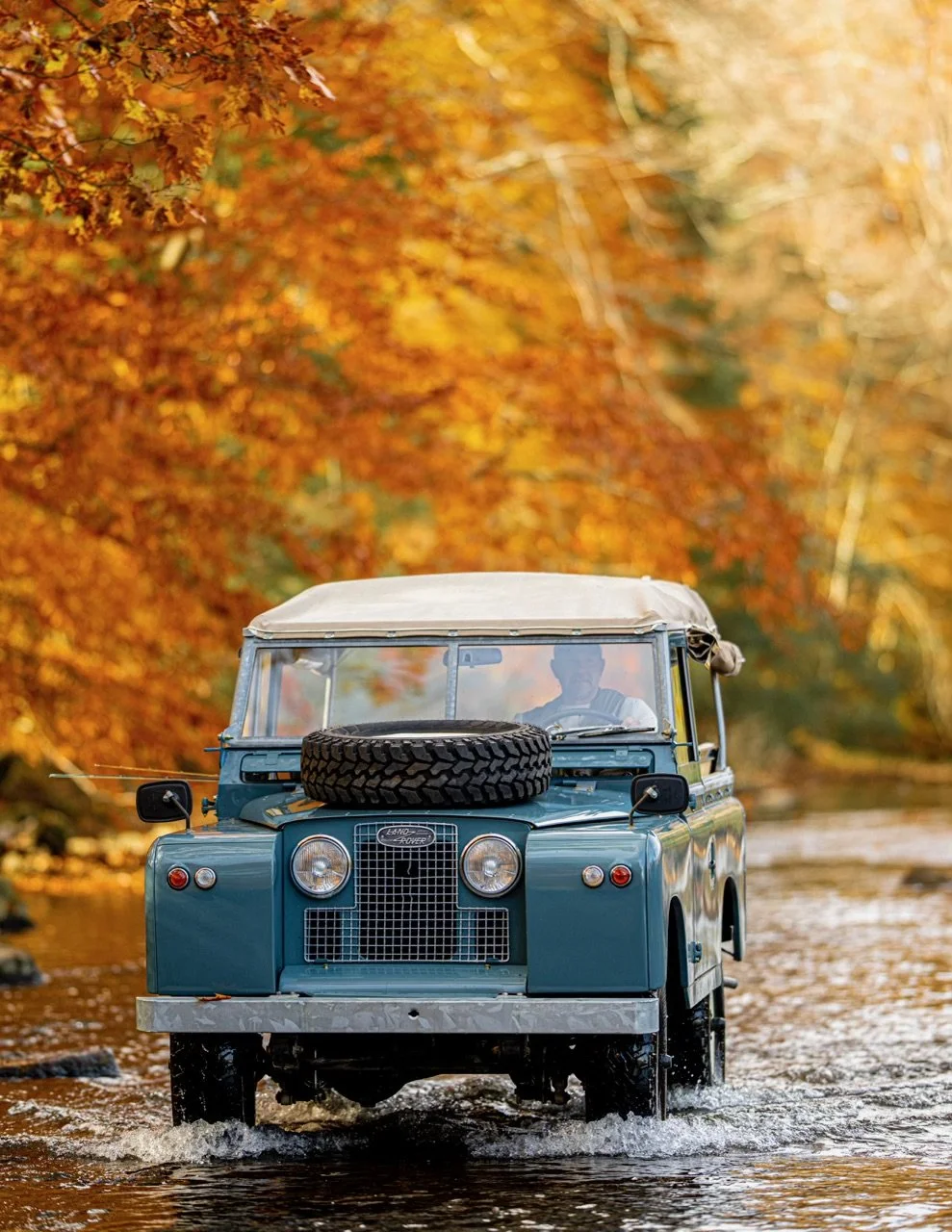 A vintage Land Rover driving through a shallow river surrounded by vibrant orange and yellow autumn trees.