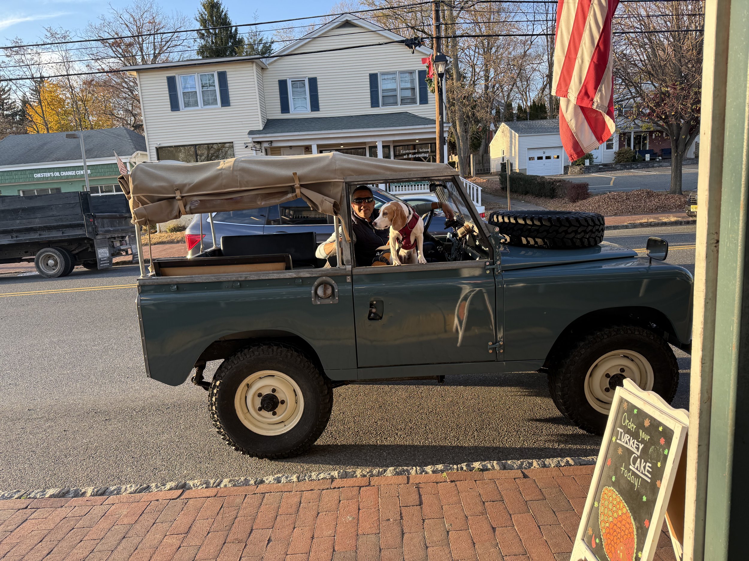 A man and a dog sitting in a small gray jeep with a beige canopy, parked on the street. An American flag is visible, and there are buildings, trees, and a sign for Chester's Oil Change Center in the background.