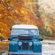 A blue vintage Land Rover driving on a road surrounded by autumn trees with orange and yellow leaves.