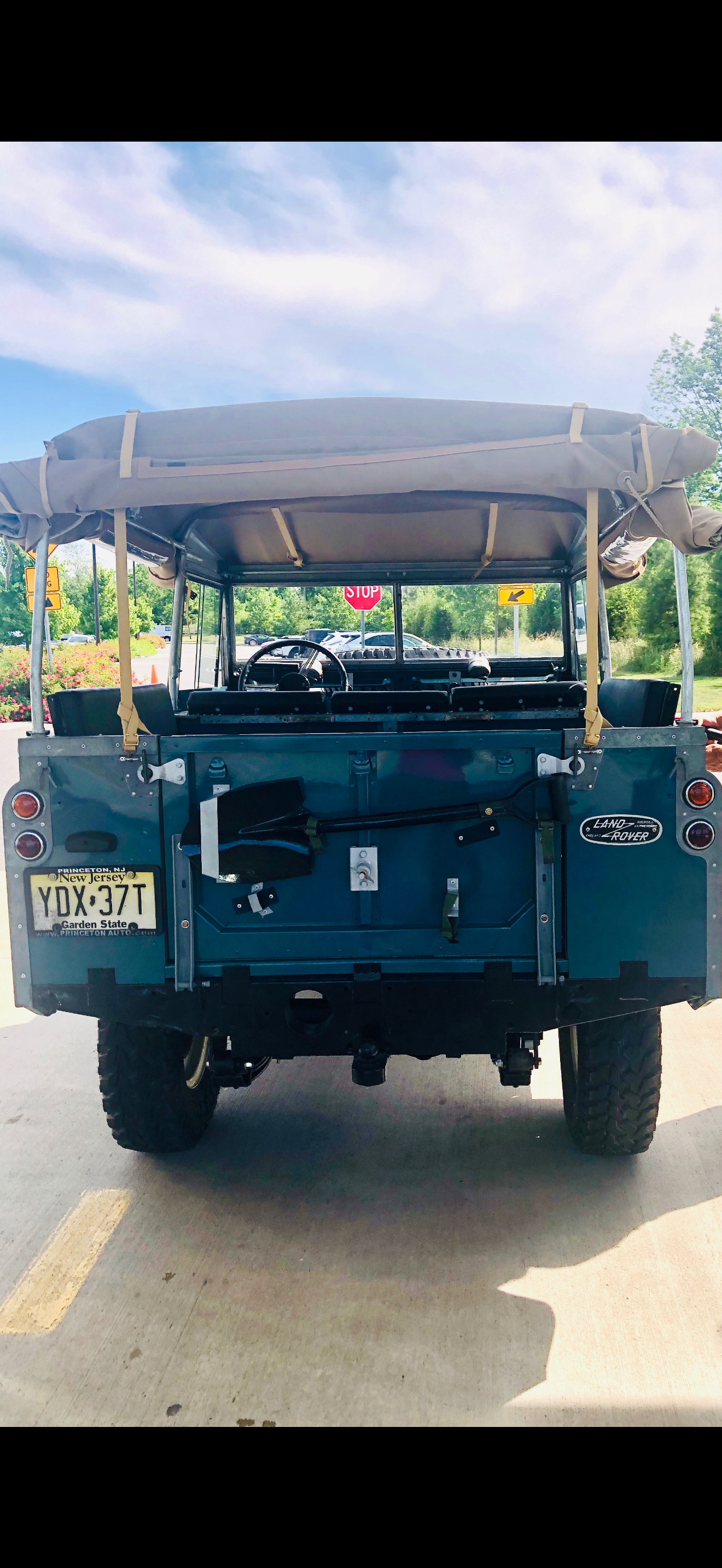 Rear view of a vintage blue Land Rover with a beige canvas top, parked in a lot with greenery and a stop sign in the background.