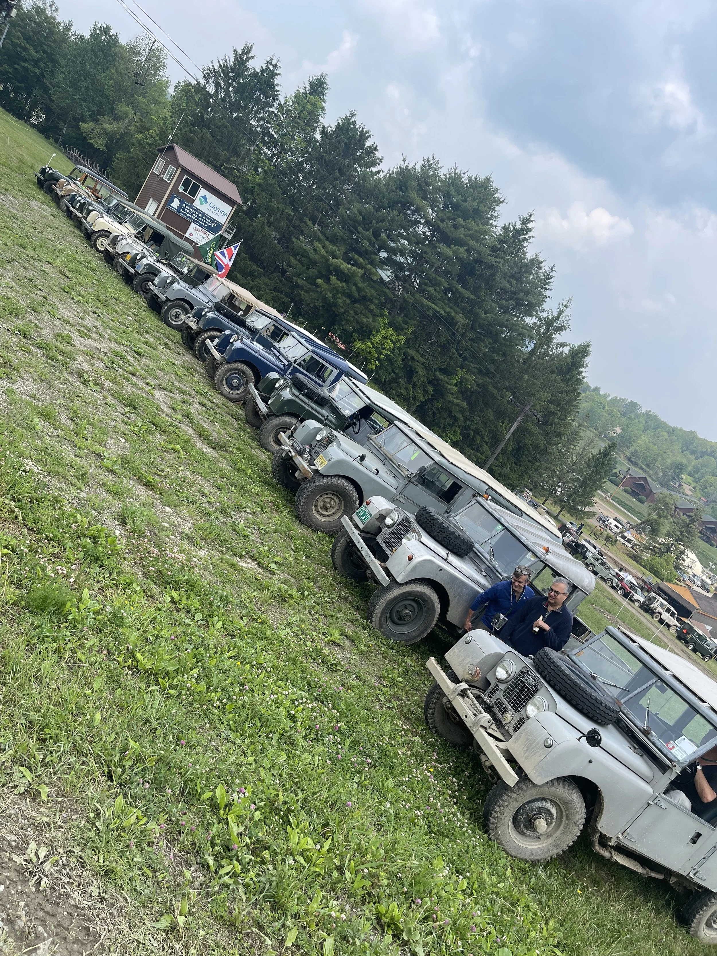 Line of vintage off-road vehicles parked on a grassy hillside, with two men standing and chatting nearby, under a partly cloudy sky with green trees and buildings in the background.