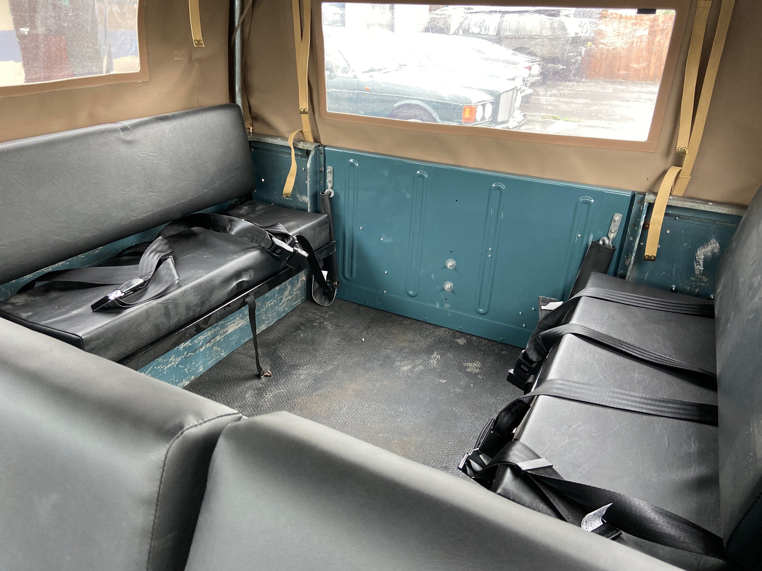Interior of a vintage vehicle with black leather seats, beige fabric-covered walls, and large windows showing an outdoor parking lot with cars and a wooden fence.