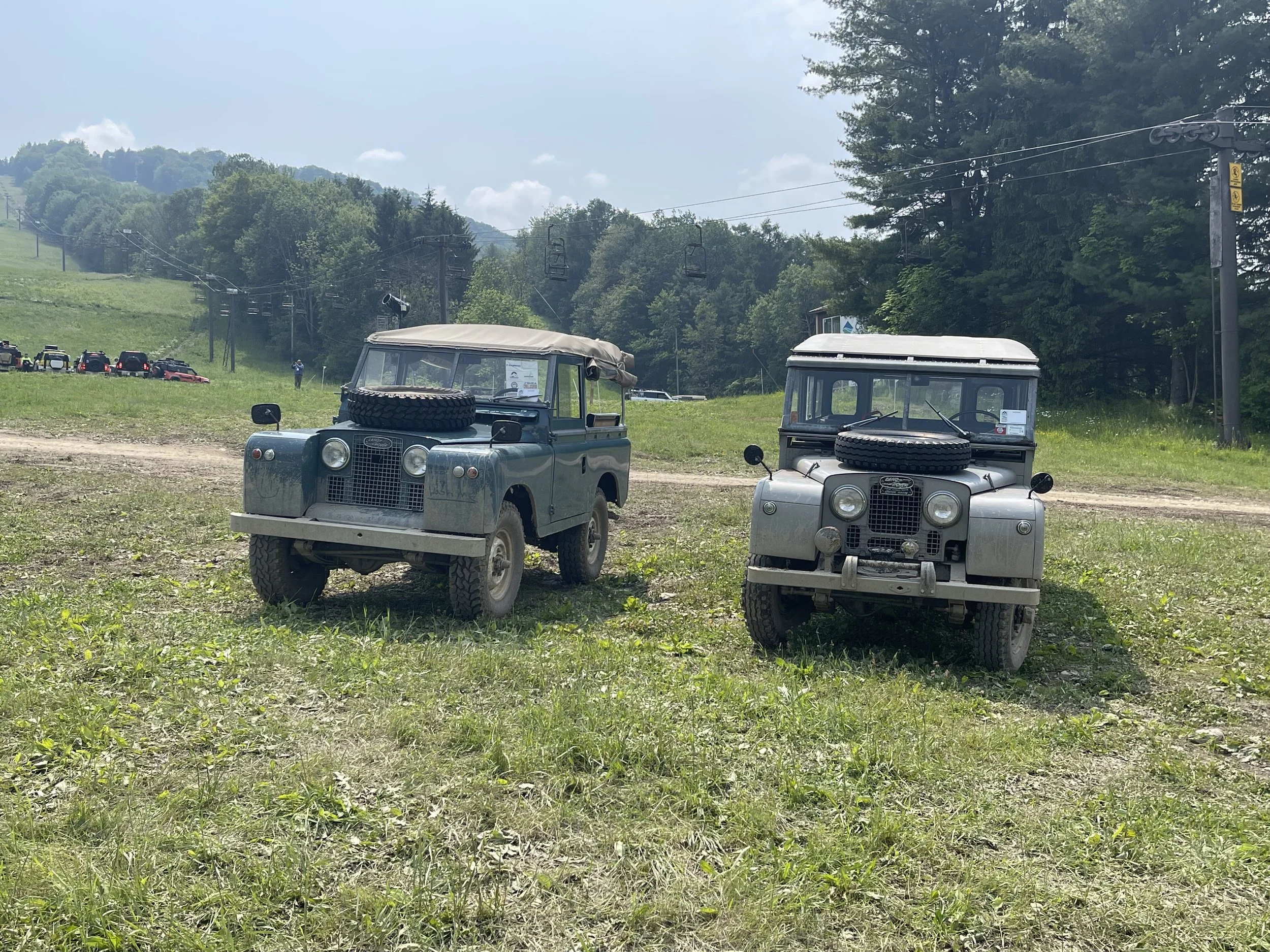 Two vintage off-road utility vehicles with spare tires on the front, parked on a grassy area near a ski lift or chairlift. In the background, there are trees, a line of cars, and a person taking a photo, with a backdrop of wooded hills under a partly