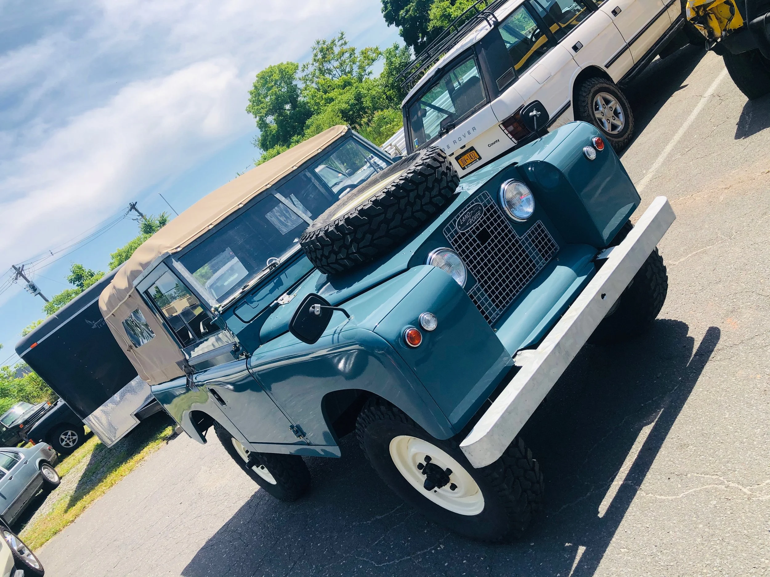 A light blue vintage Land Rover Defender parked next to a white Land Rover Discovery in a parking lot with green trees and a partly cloudy sky in the background.