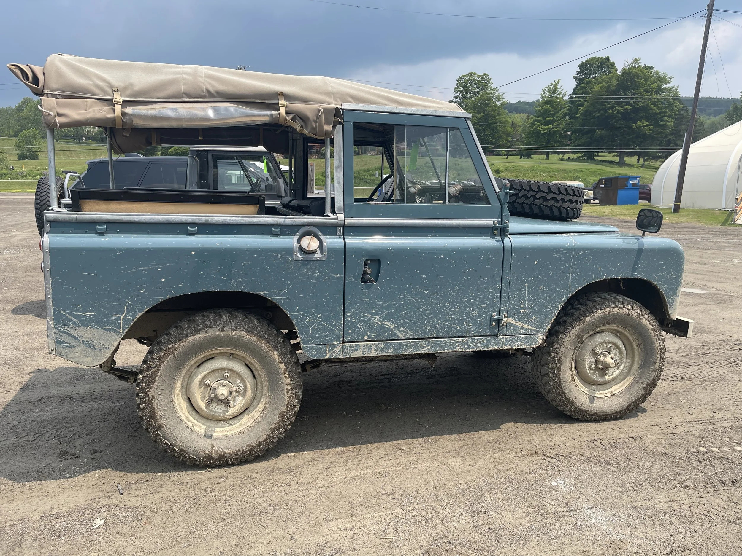 A blue vintage off-road vehicle with a beige canopy covering the back, parked on a dirt surface under an overcast sky, with a scenic green landscape and utility poles in the background.