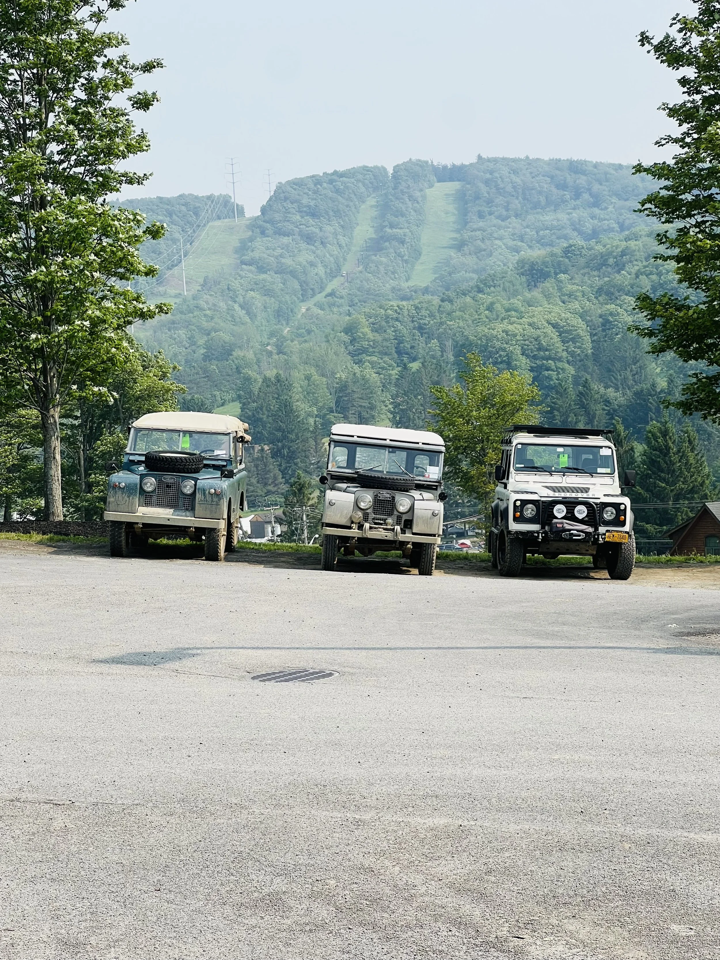 Three vintage Land Rover vehicles parked on a flat road with a wooded mountain and power lines in the background.