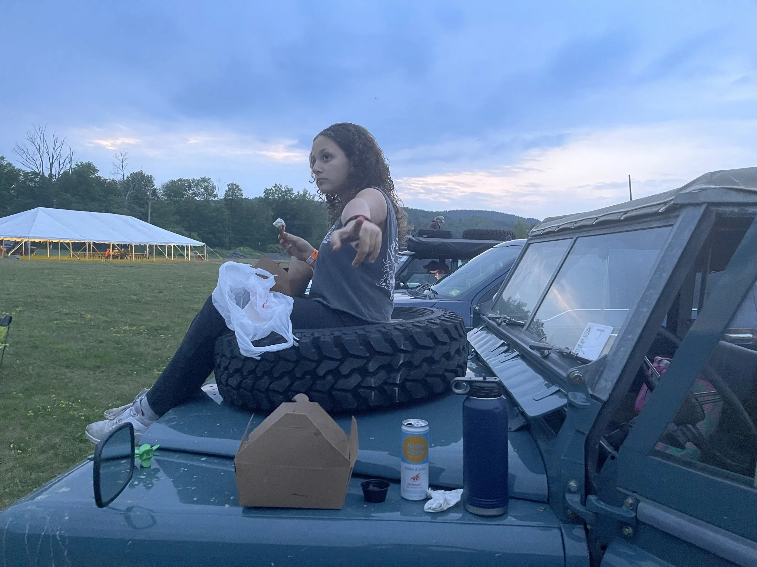 A young woman with curly hair sitting on the hood of a vehicle at an outdoor event during dusk, surrounded by other vehicles, a large white tent, and a landscape with trees and hills.