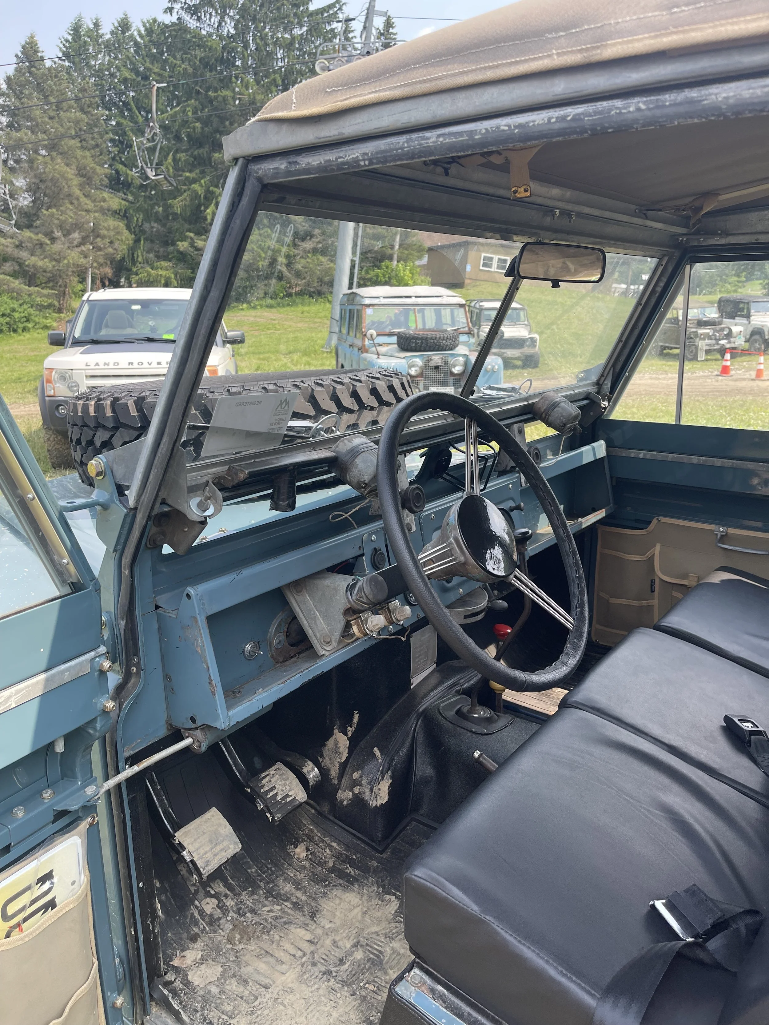 Inside a vintage off-road vehicle with a black bench seat, gear shift, and steering wheel, overlooking a grassy field with parked Land Rover and other vehicles, trees, and power lines in the background.