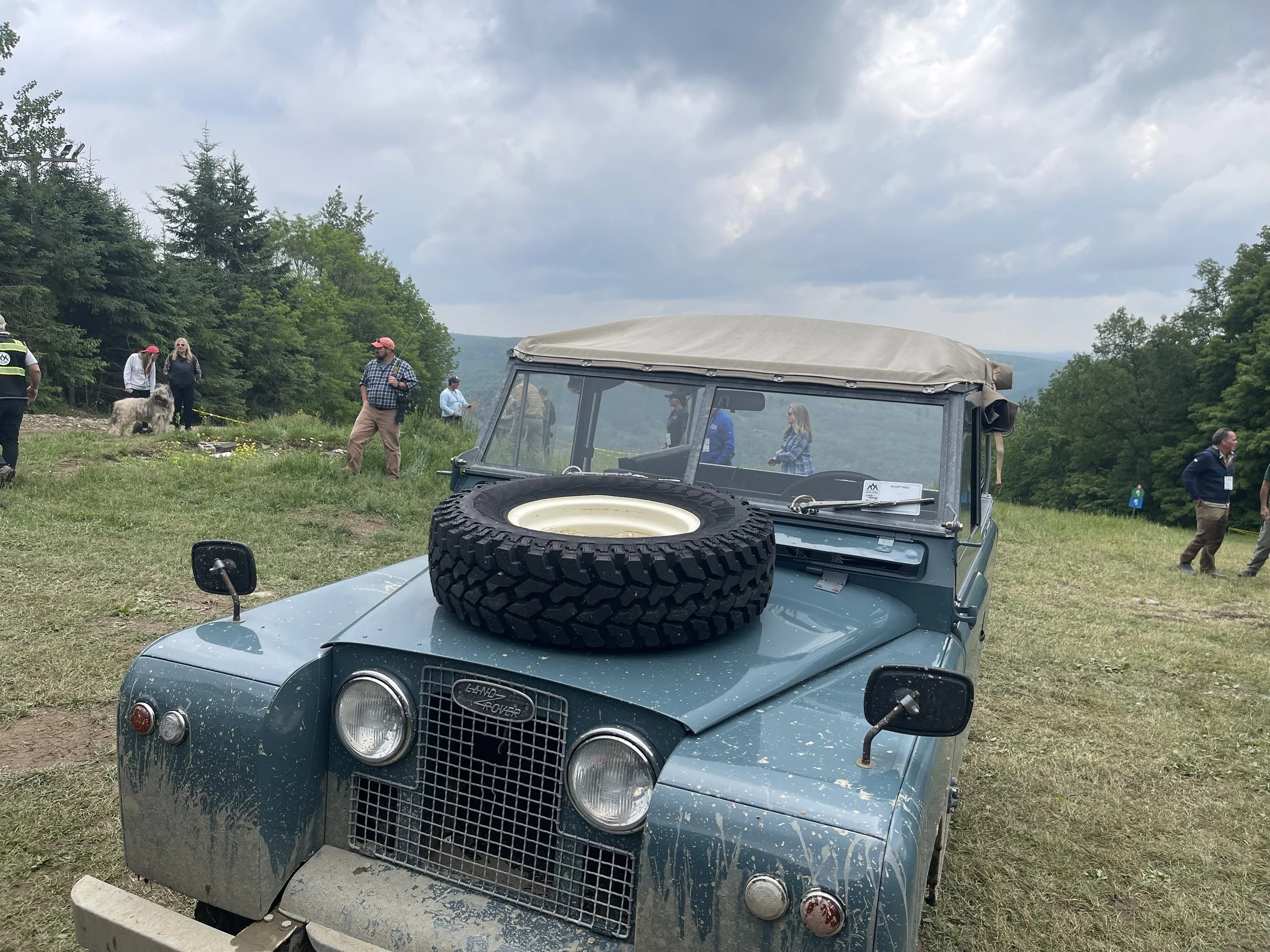 A vintage blue Land Rover with a tire mounted on the hood parked on grassy terrain during an outdoor event with people, trees, and cloudy sky in the background.