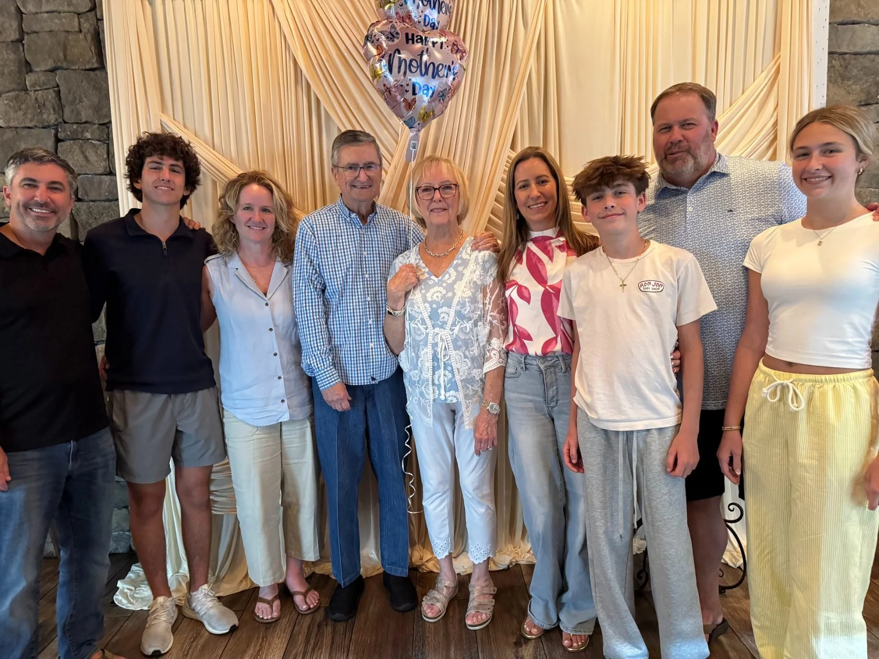 A group of ten people, including elderly, middle-aged, and young adults, stand together indoors in front of a beige curtain backdrop with balloon decorations. Some are smiling at the camera, and there is a sign on one balloon saying 'Happy Mother’s D