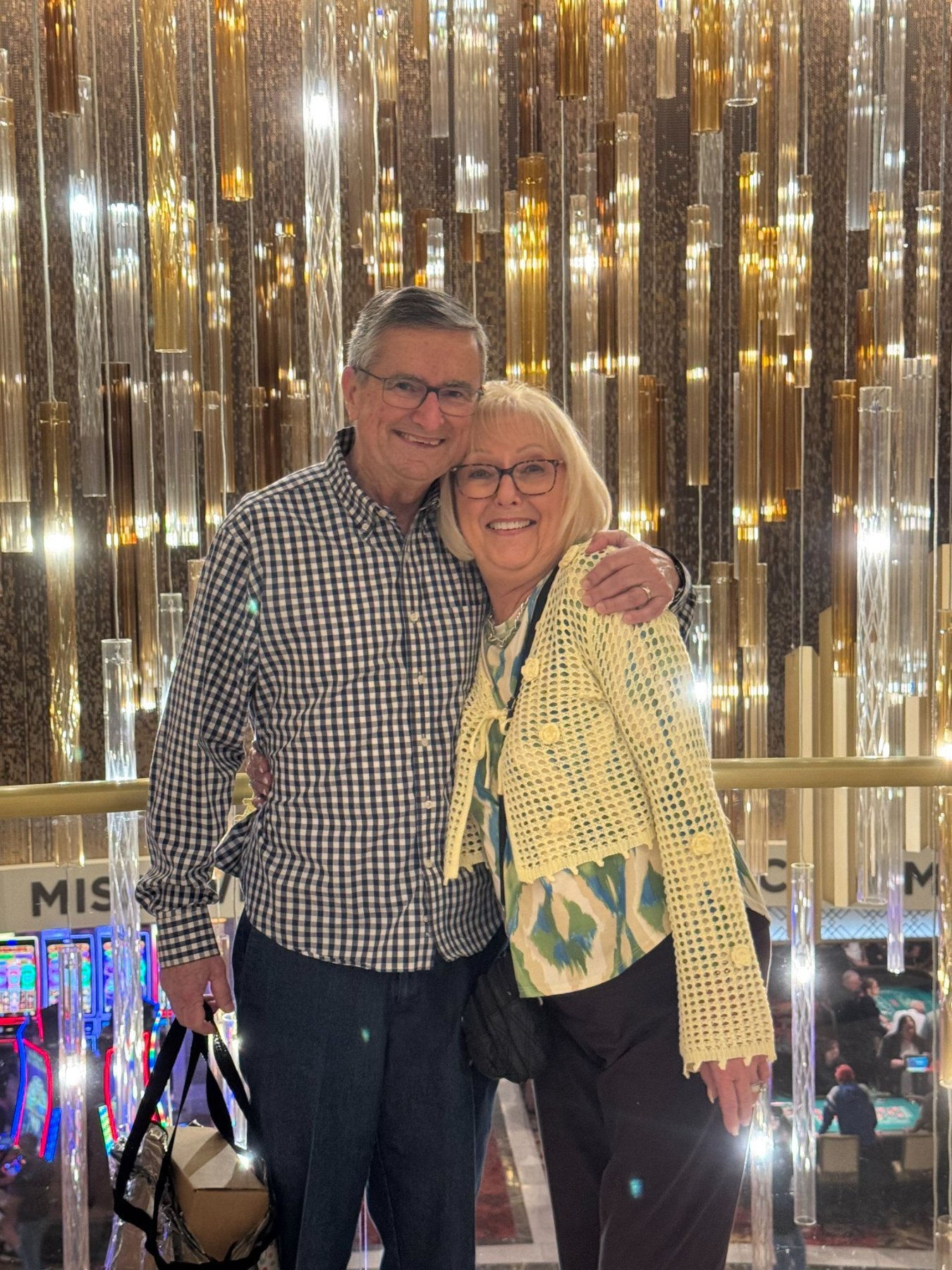 A smiling older man and woman standing together in front of a decorative, illuminated backdrop with vertical gold and clear glass tubes, inside a lively venue that appears to be a casino or entertainment space.