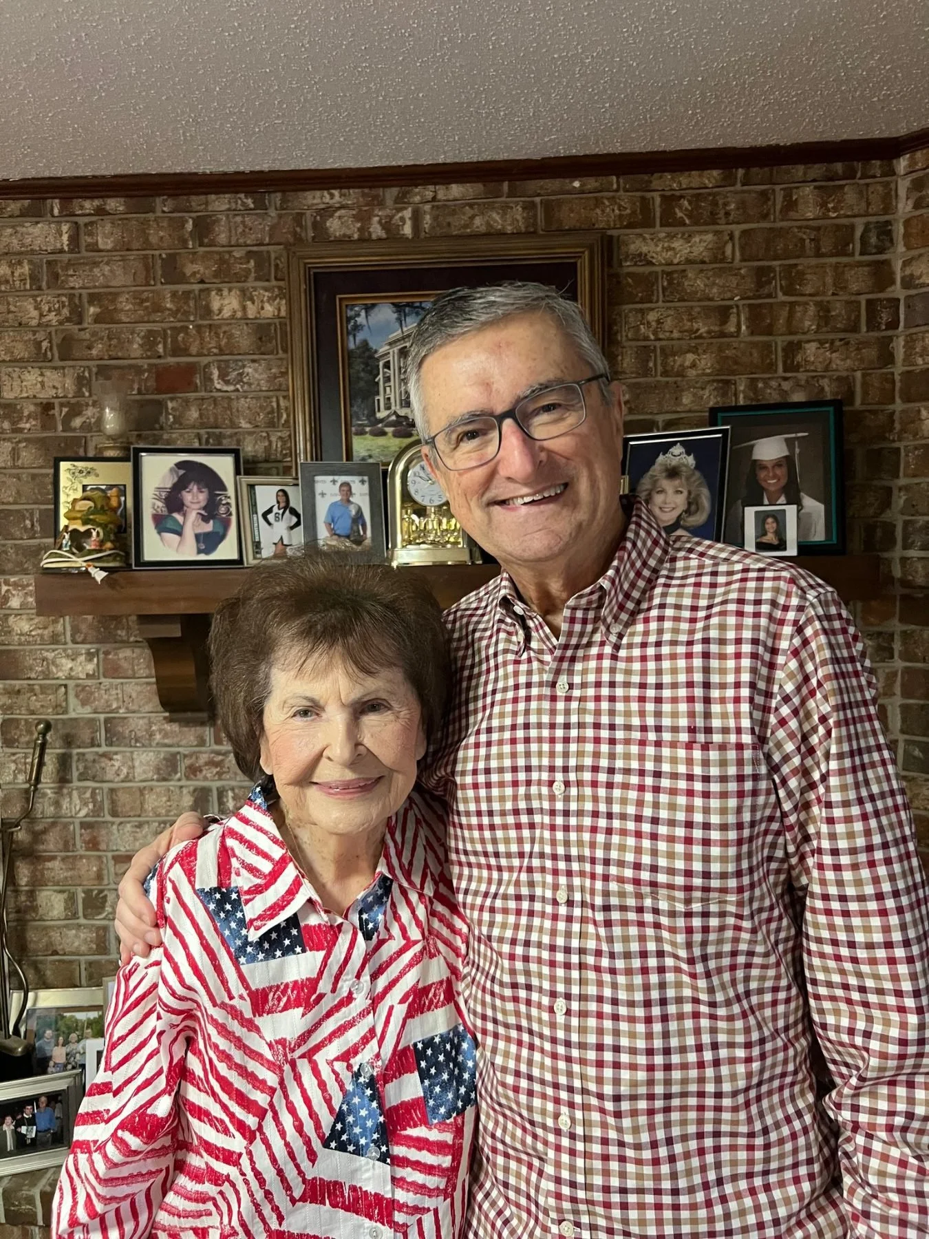 An elderly woman and an elderly man smiling and posing together in front of a brick wall with framed photographs and a mantelpiece behind them.