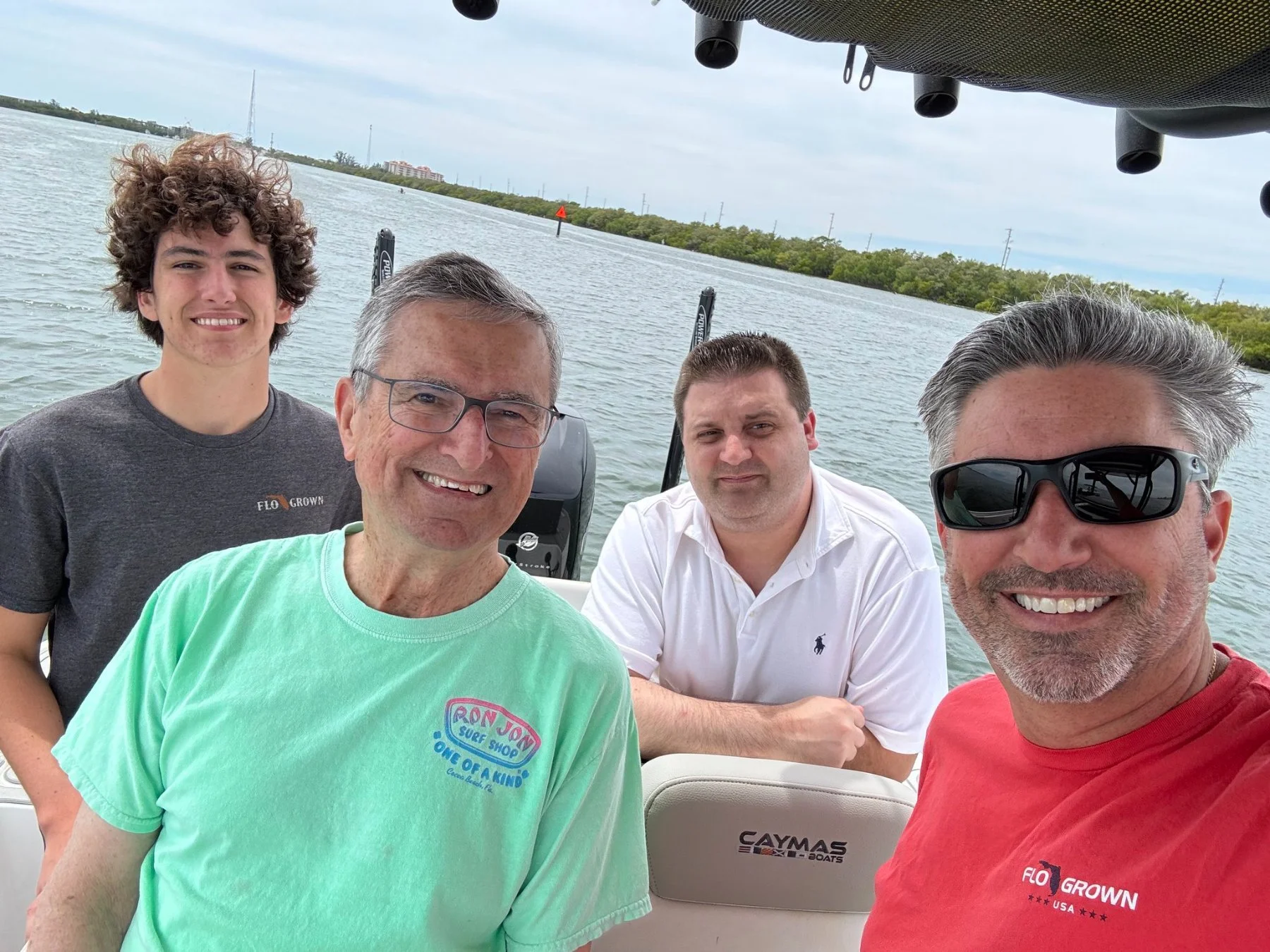 Four men taking a selfie on a boat with water and trees in the background. They are smiling and appear to be enjoying a day on the water.