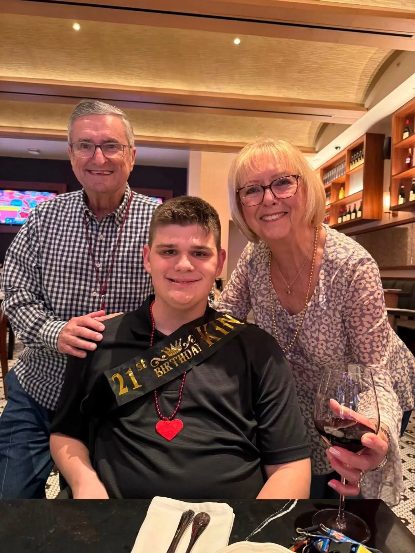 A young man celebrating his 21st birthday at a restaurant with his family, wearing a black shirt with a "21st Birthday" sash, a red heart necklace, and a smile. Two older adults, a man and a woman, stand behind him, smiling. The man is wearing glasse
