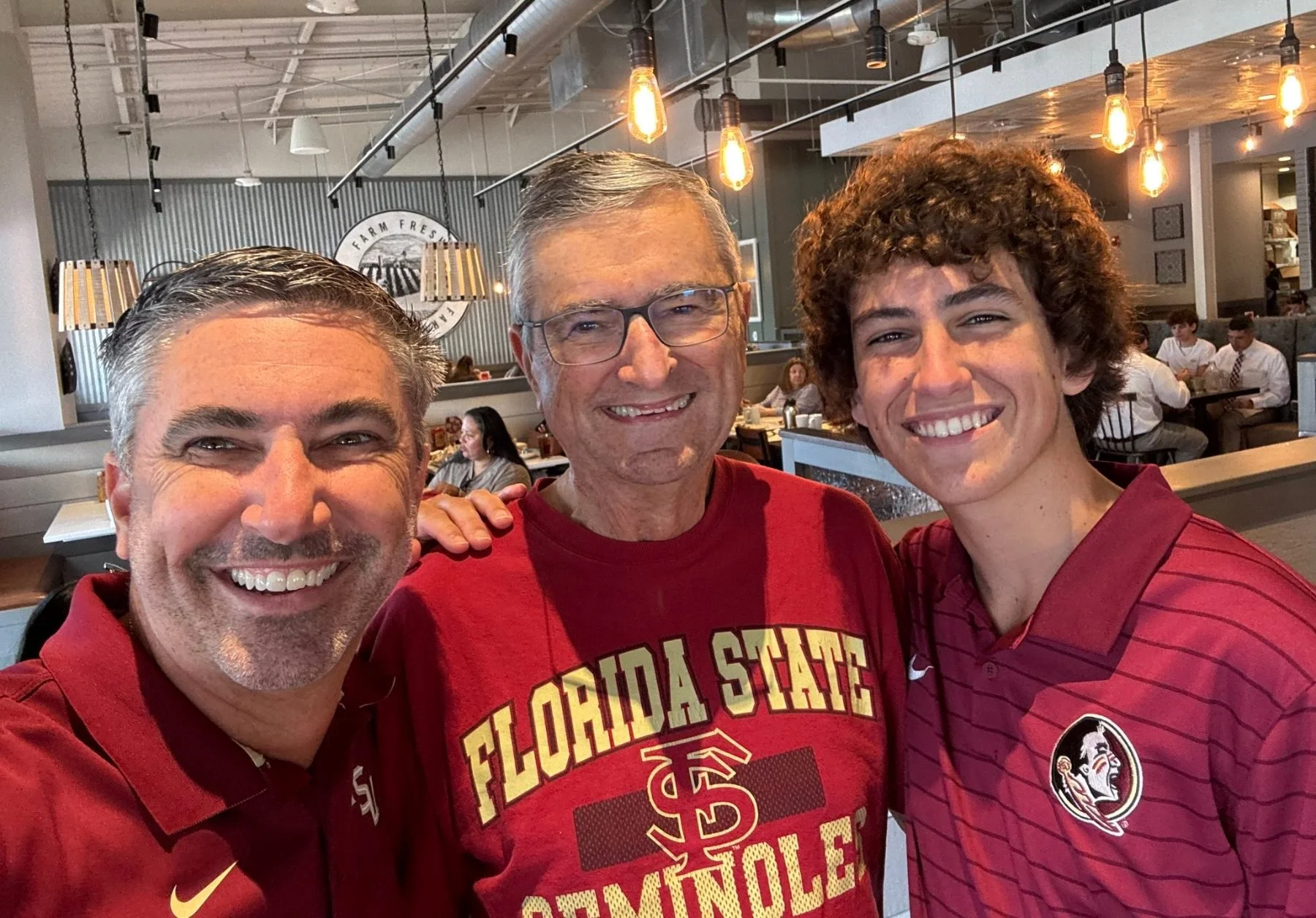 Three men smiling and taking a selfie in a restaurant, with a background of other diners and warm lighting.