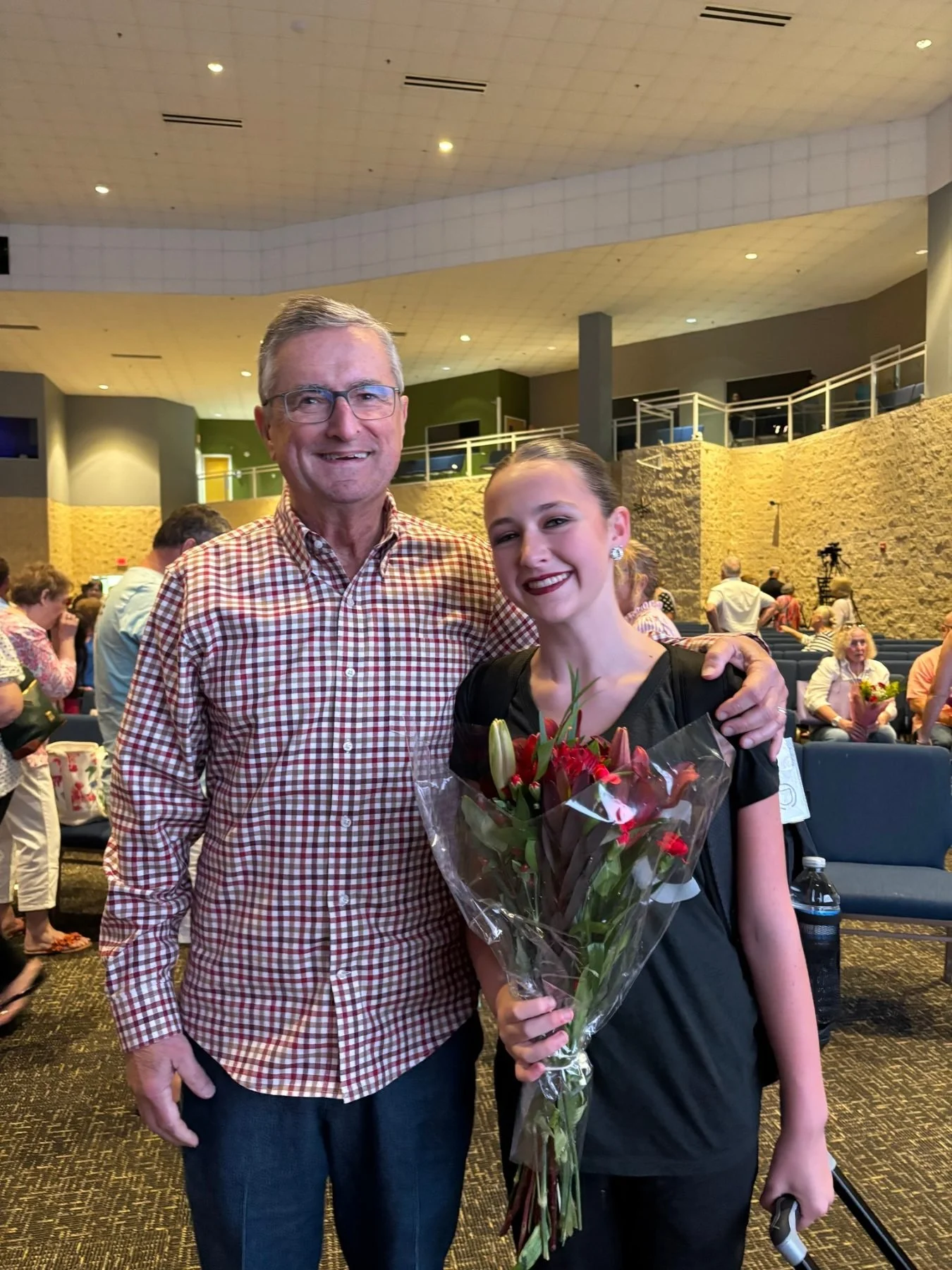 An older man and a young woman standing together in a large, indoor event space. The woman is holding a bouquet of flowers and smiling, with the man smiling beside her. In the background, people are seated and standing, and there is a camera on a tri