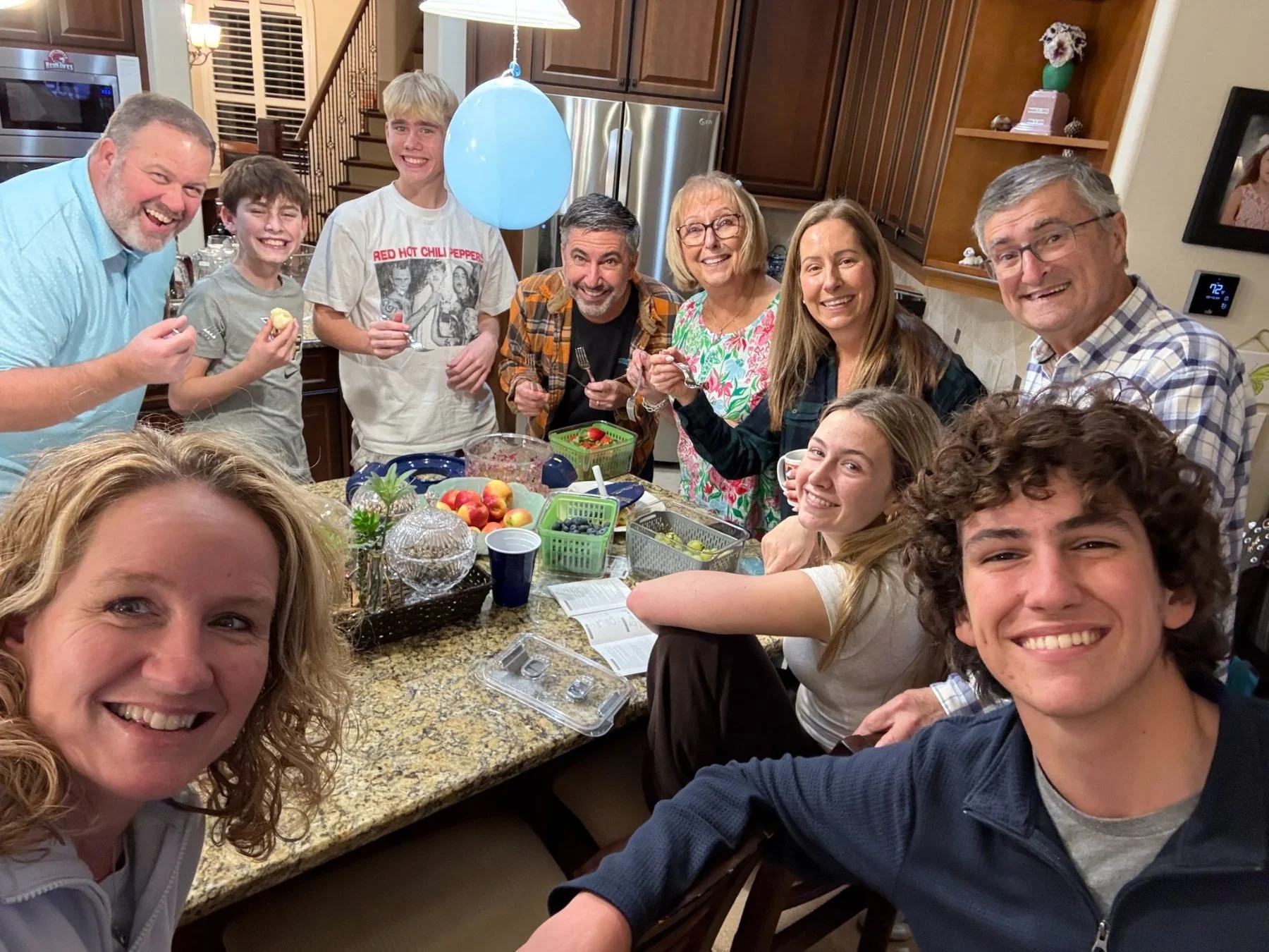 A group of people gathered around a kitchen table, smiling and celebrating. The table has fruit bowls and snacks, and the group appears to be enjoying a joyful moment together.