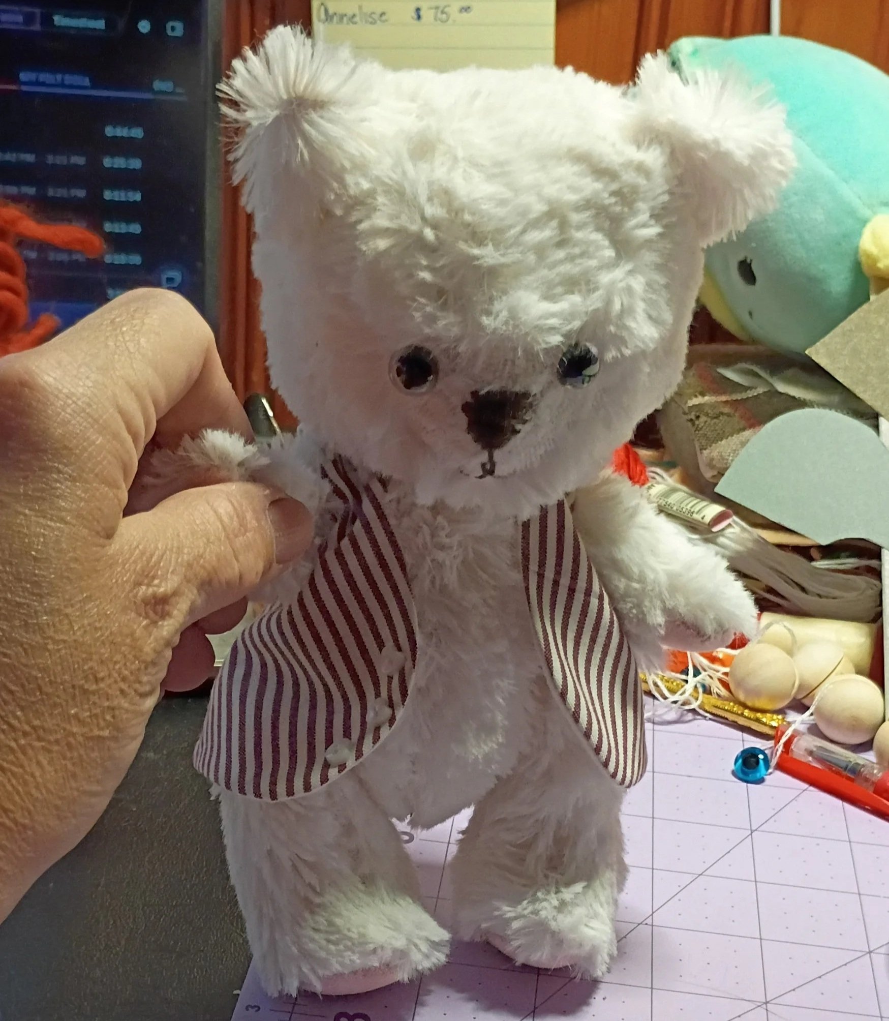 A person in a striped shirt is holding a white fluffy teddy bear with black eyes and nose, with a background of toys and a yellow sign with some writing.