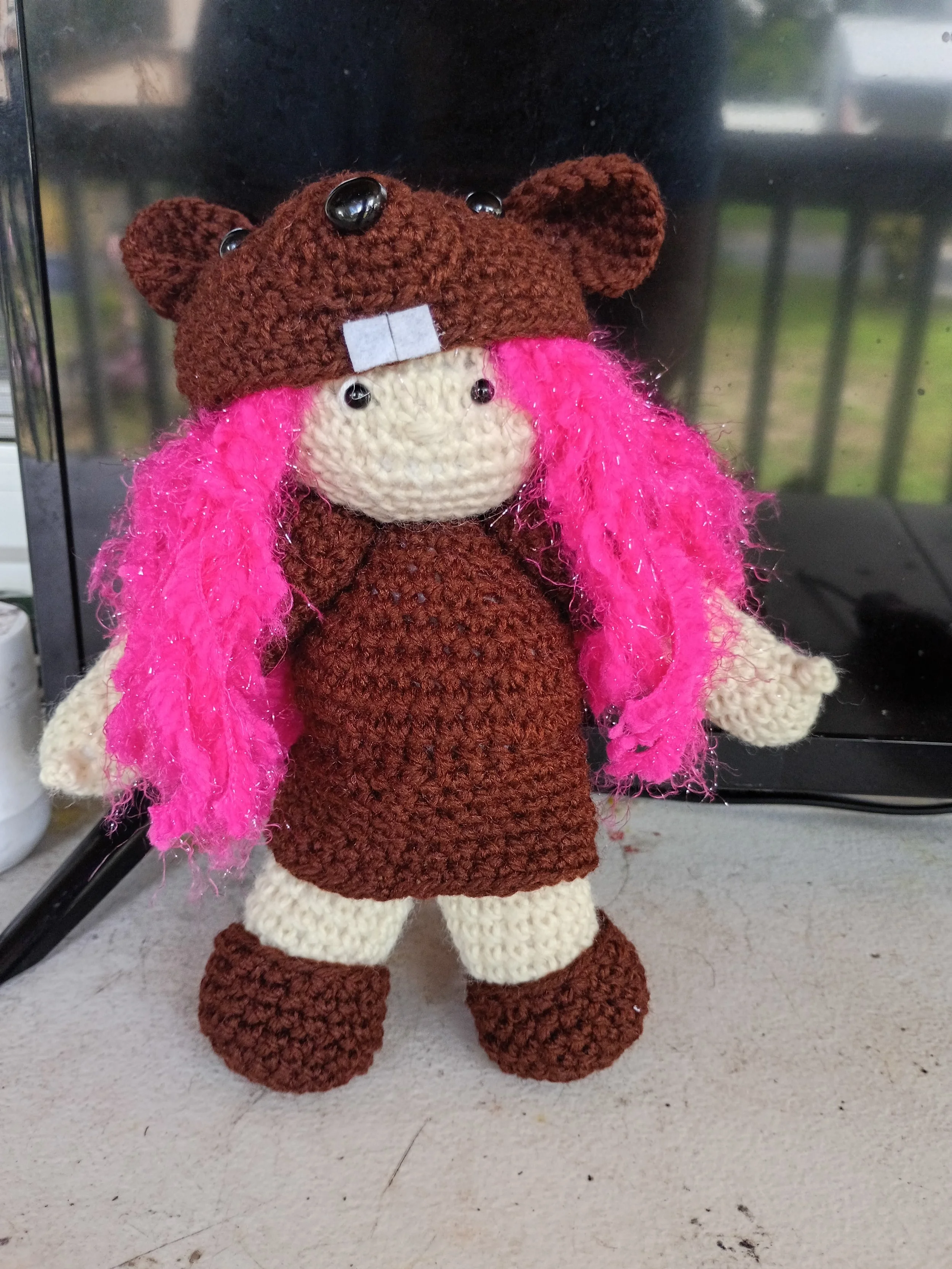 A crocheted stuffed animal resembling a beaver with a small gray bow on its head, large black eyes, a beige face, pink hair, and brown and beige limbs, standing on a beige surface against a black background.